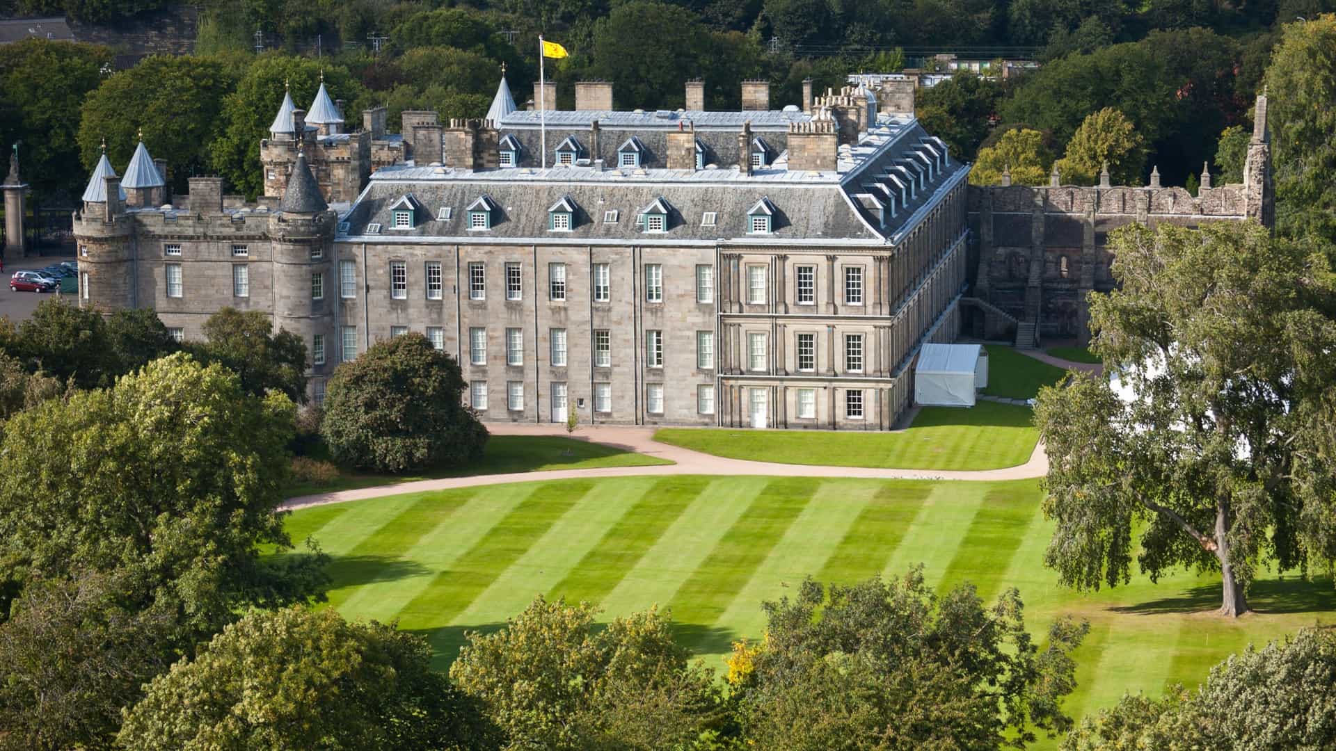  The Palace of Holyroodhouse, the official royal residence in Edinburgh, Scotland. The image shows the palace's grand facade, a mix of historic and Baroque architecture, with its prominent towers and stone courtyard. The setting includes the palace's grounds and the lush green landscape of Holyrood Park, with the dramatic hills of Arthur's Seat visible in the background.