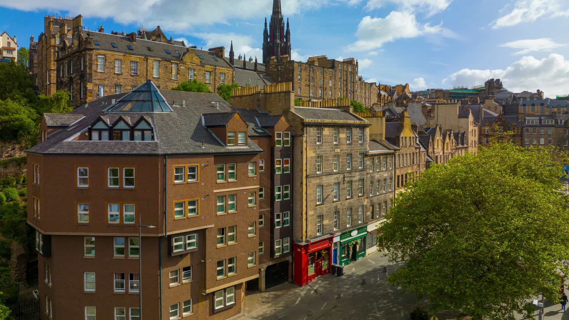 An aerial view of Edinburgh's historic Old Town, focusing on the intersection of the Grassmarket area and the iconic Royal Mile. Dominating the skyline is the Gothic Revival spire of the Tolbooth Kirk (The Hub), which dramatically pierces the sky above the cluster of historic, tightly packed stone buildings. The image captures the medieval street layout and the area's designation as a UNESCO World Heritage Site.