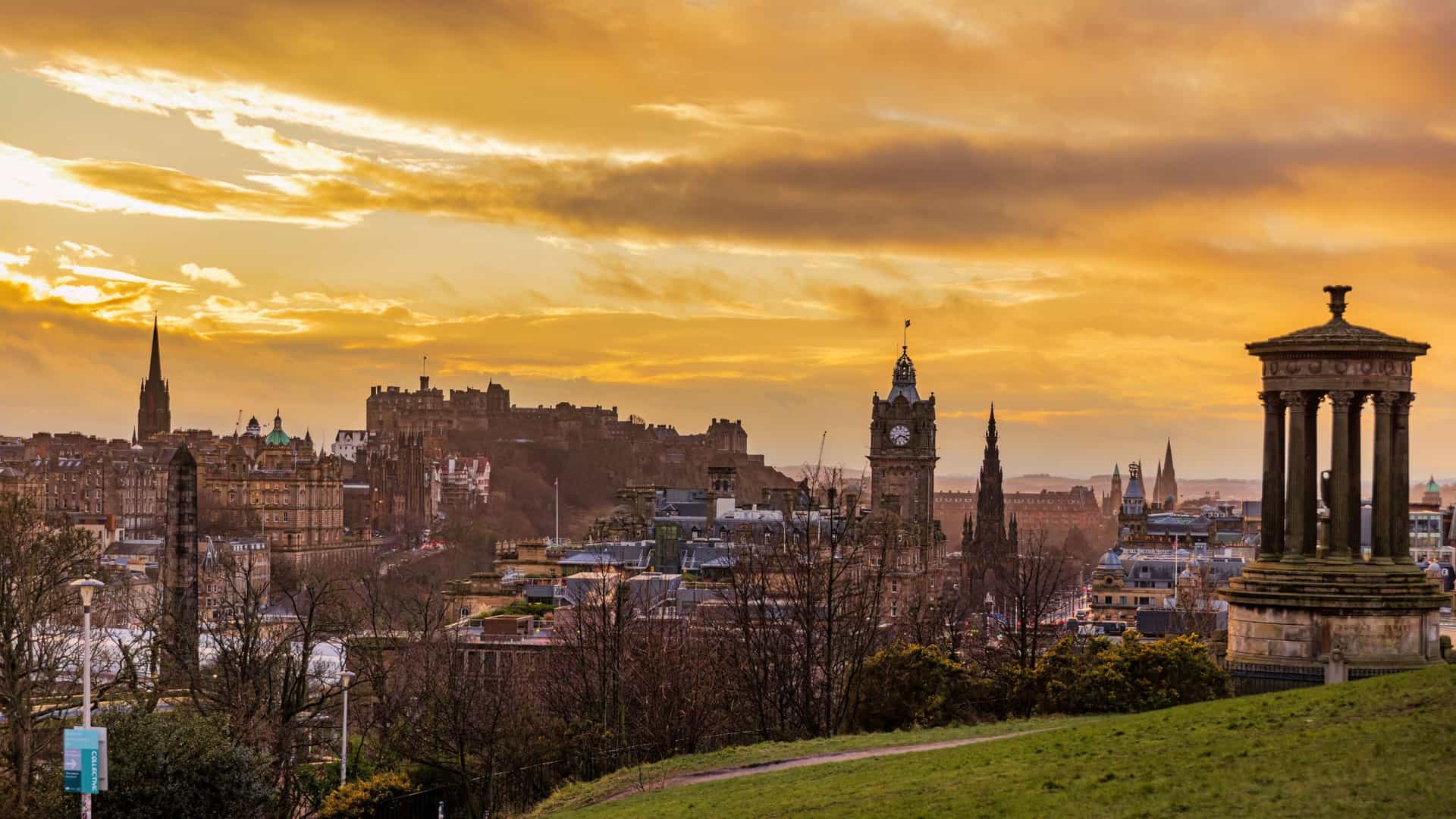 A scenic view of the city of Edinburgh from the top of Calton Hill. The image shows the National Monument, a structure of large columns resembling the Parthenon, in the foreground. In the distance, the cityscape of Edinburgh unfolds, with buildings and spires visible under a wide sky.