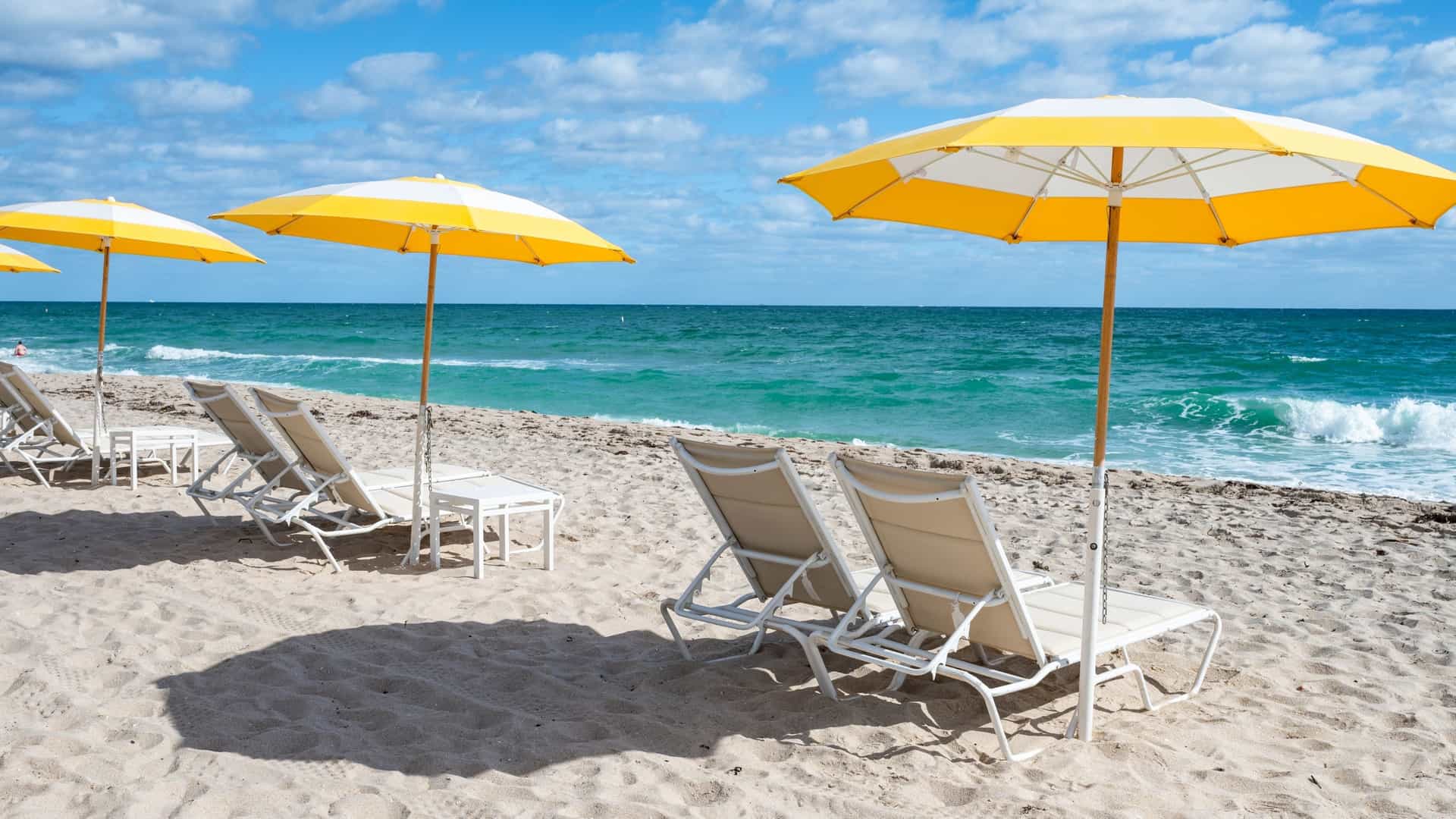 A bright and sunny view of Fort Lauderdale Beach. The foreground is filled with rows of colorful beach chairs and large umbrellas, indicating a rental setup. Beyond the beach, the turquoise-blue ocean stretches out to the horizon under a clear sky. The scene conveys a sense of relaxation and leisure on a classic Florida beach.