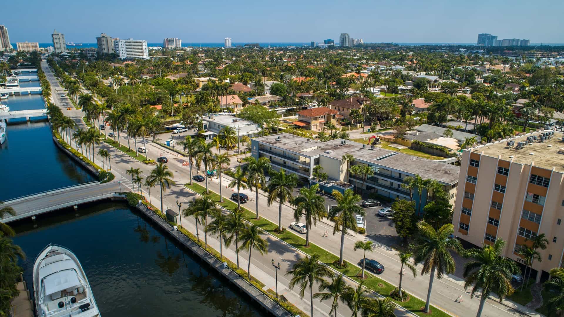 An aerial drone shot of Las Olas Boulevard in Fort Lauderdale, Florida. The image captures the urban landscape, with the main boulevard running through a developed area of high-rise buildings, residential neighborhoods, and a network of canals. The canals are lined with docks and feature numerous boats and yachts. The image showcases the city's blend of modern architecture and waterfront living.