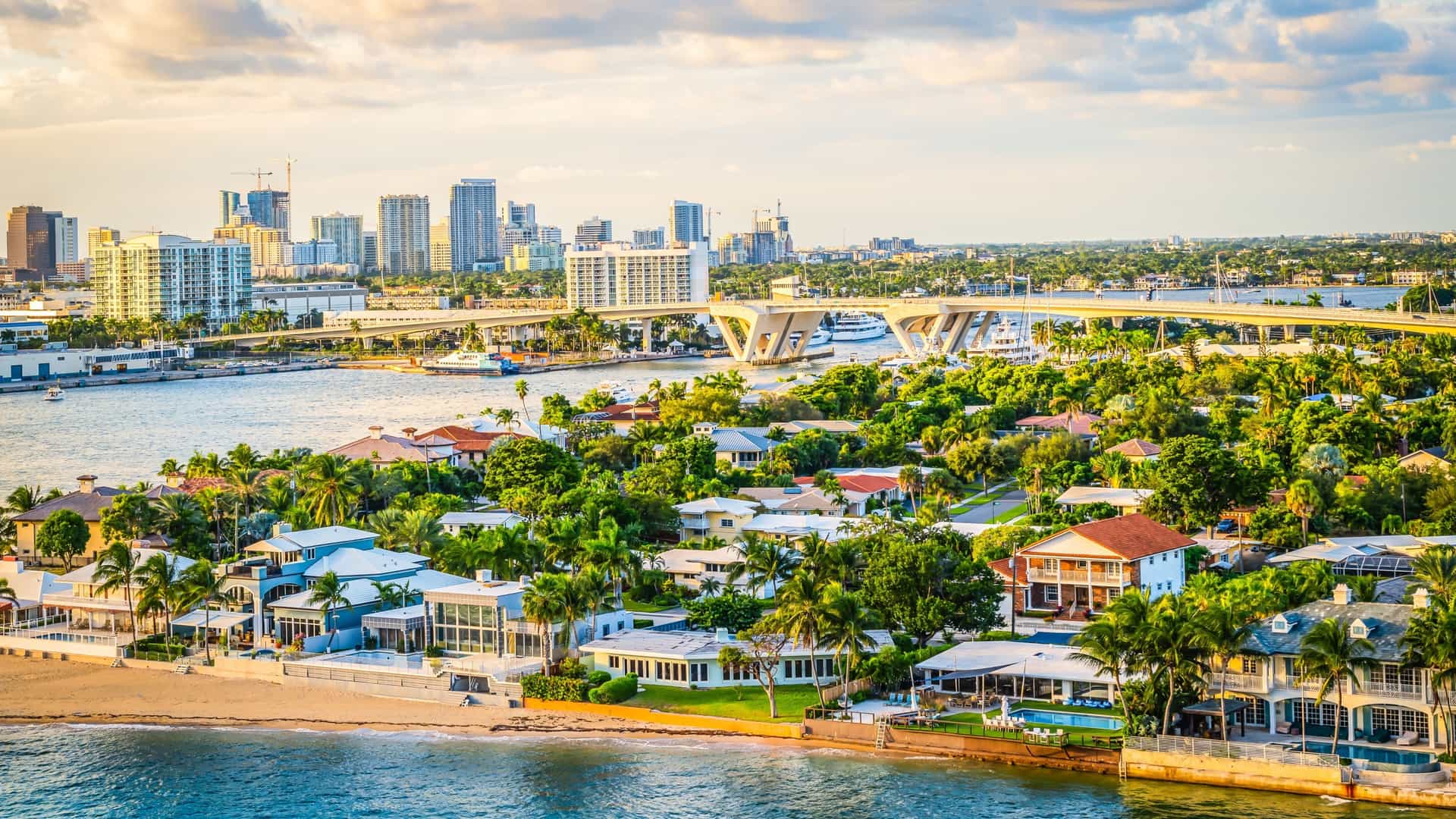 A scenic view of the Intracoastal Waterway in Fort Lauderdale, Florida. The waterway is lined with a variety of luxurious, multi-story waterfront houses, each with a private dock. Several boats and yachts are moored in the water, highlighting the area's popular boating lifestyle. The image captures the upscale residential architecture and the beautiful canal system of the area.