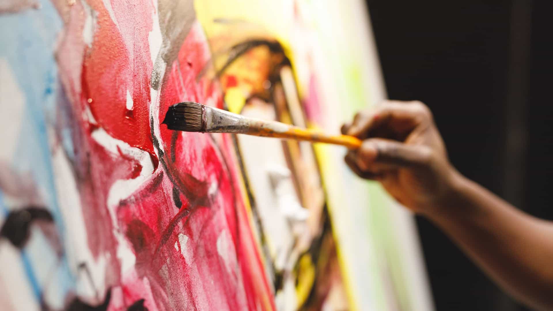 A close-up shot of a male painter at work in the NSU Art Museum studio, intensely focused as he applies paint to a canvas with a paintbrush. A colorful palette is visible in his hand.