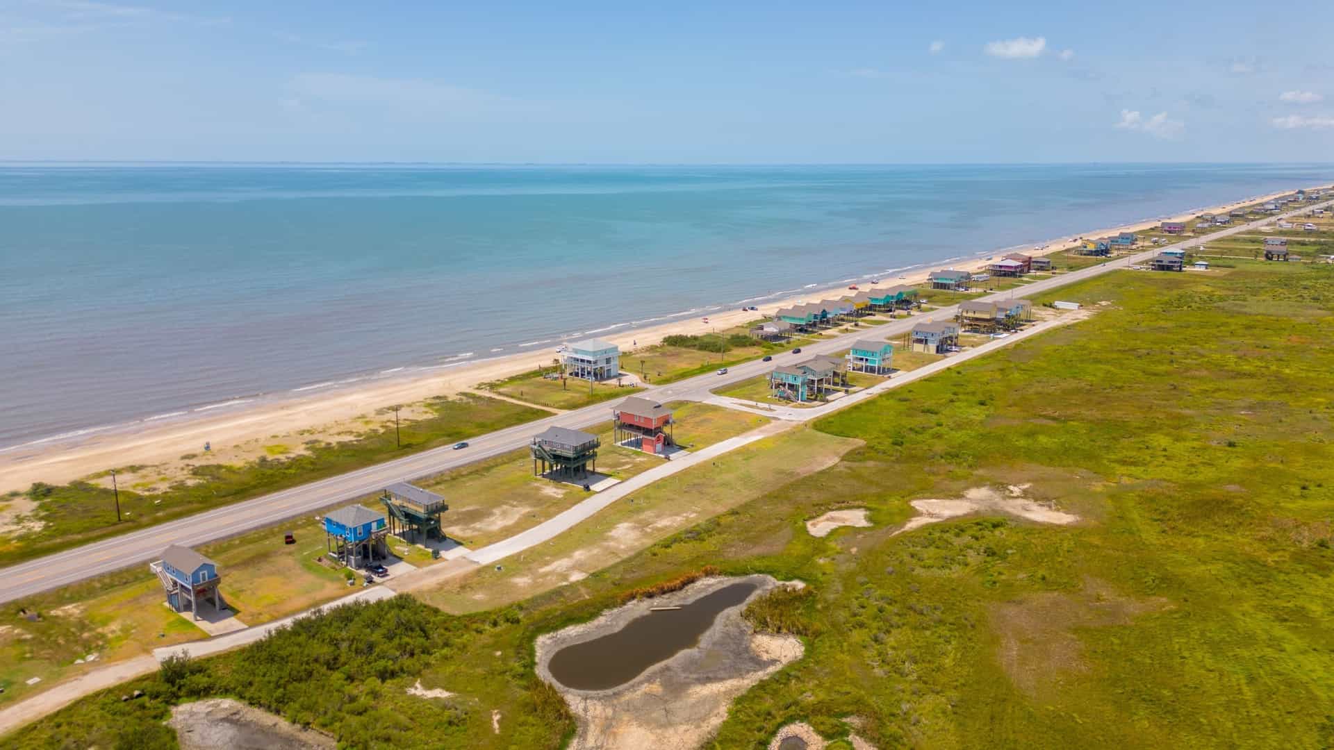 A row of beach houses built on wooden stilts stands along the coast of Port Bolivar, Texas. The elevated homes are designed to protect against flooding and overlook the sandy terrain.