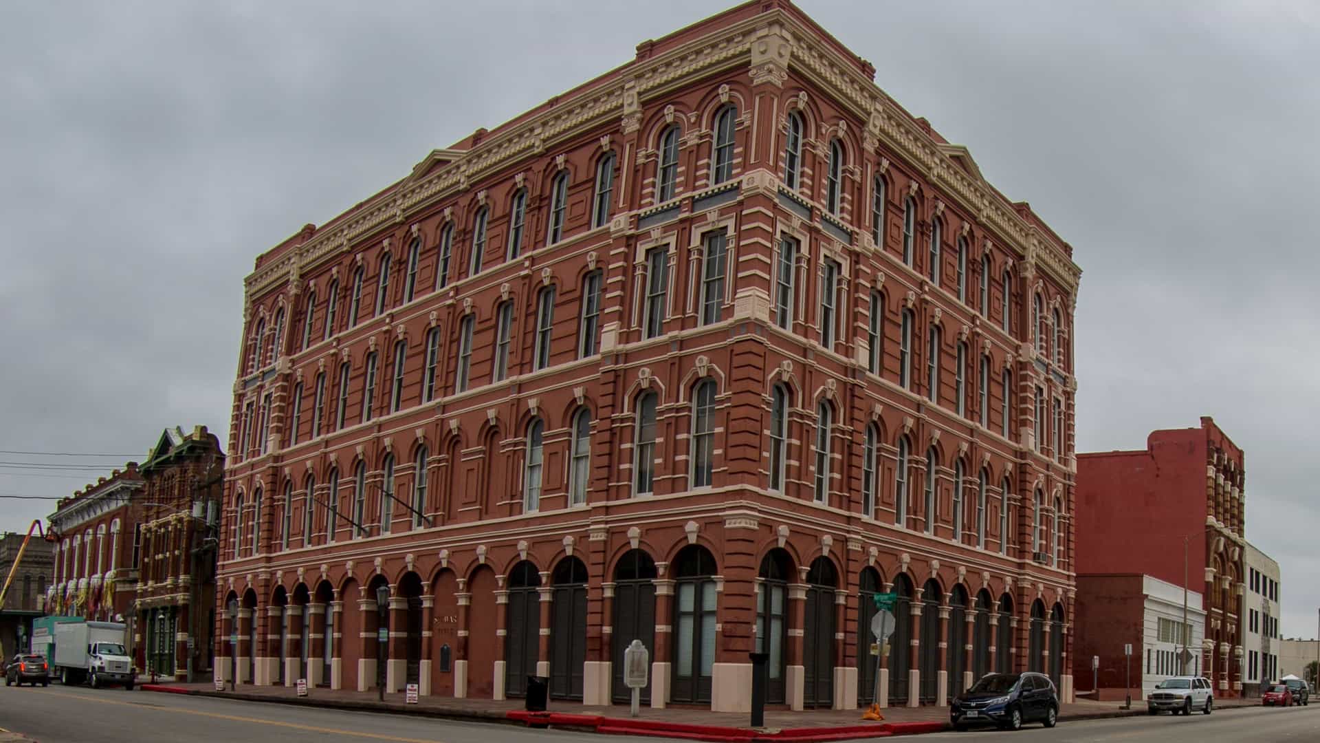 A close-up view of a multi-story Victorian commercial building with a detailed brick facade in Galveston's Strand District. The architecture features ornate windows and an antique clock mounted on the corner of the building, characteristic of the 19th-century "Wall Street of the Southwest."