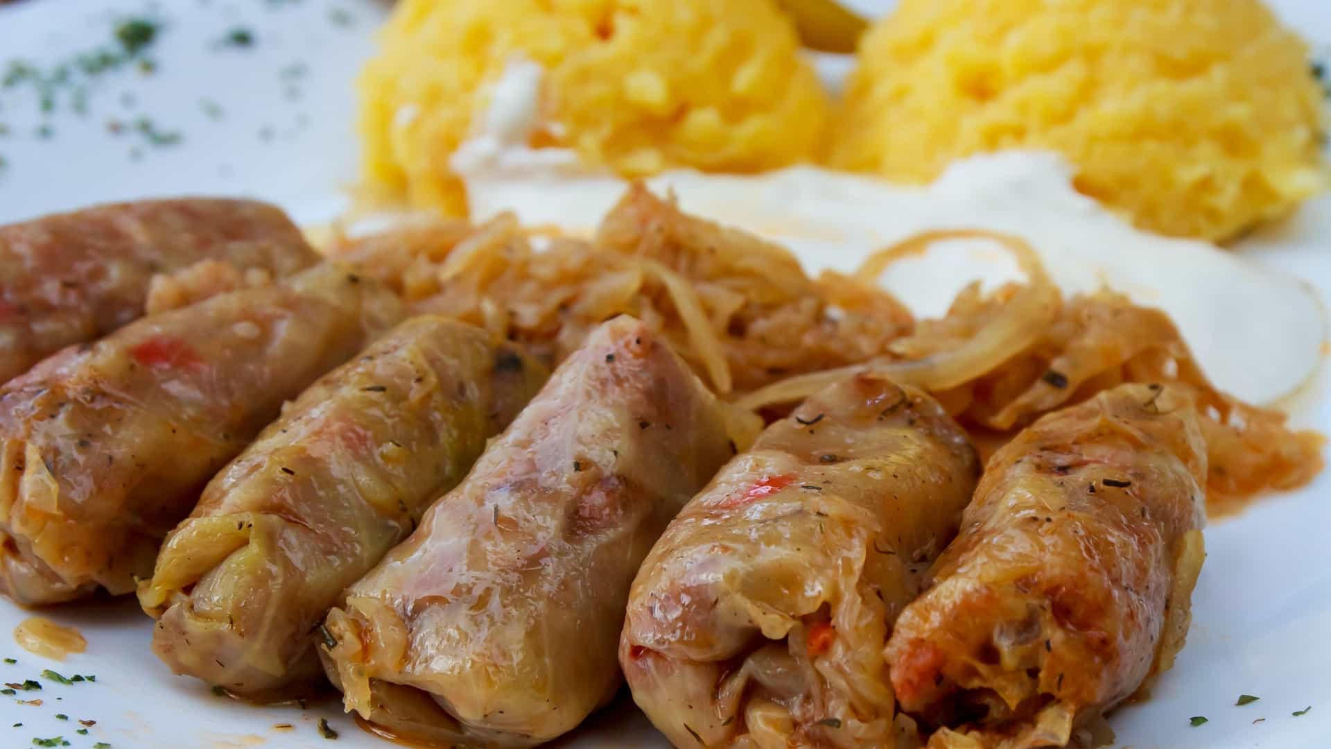 A close up shot of a plate of traditional Romanian food, "mămăligă cu sarmale." The dish consists of a thick, golden yellow cornmeal porridge (mămăligă) served alongside stuffed cabbage rolls (sarmale) topped with a spoonful of sour cream.