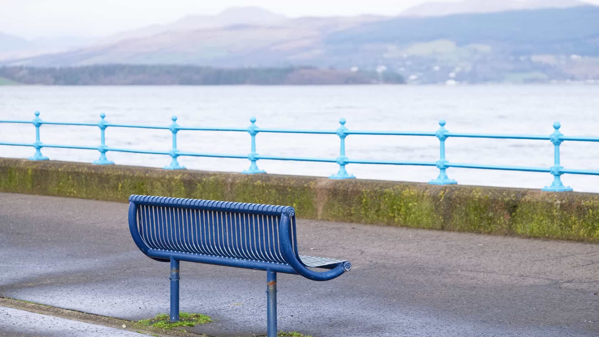 A solitary, modern, blue metal bench is situated on the paved walkway of the Greenock Esplanade Park in Scotland. The bench faces the wide expanse of the Firth of Clyde, which is framed by a distinctive bright blue metal railing and a low, mossy stone wall. In the distance, rolling green hills rise from the opposite shore under a calm, overcast sky.