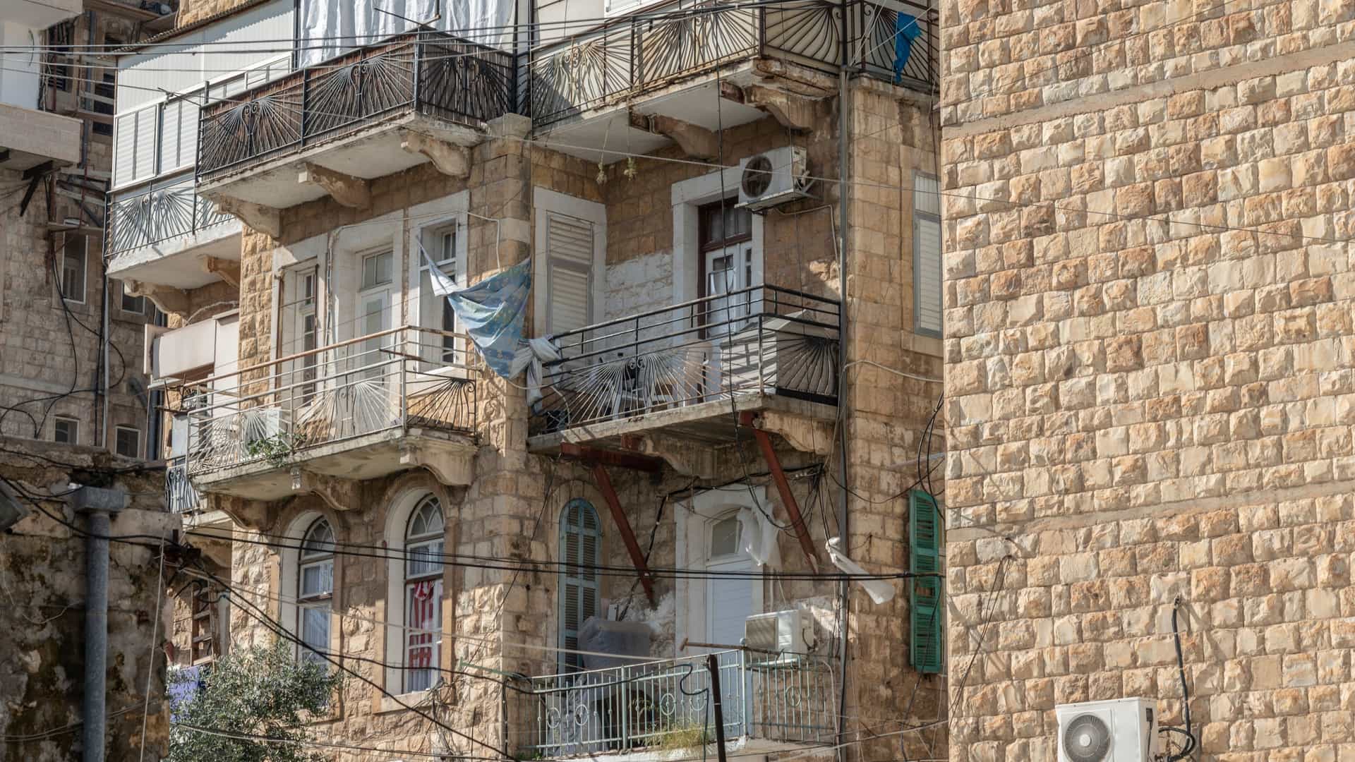 A street-level photo showcasing the architecture of the Wadi Nisnas neighborhood in the lower part of Haifa, Israel. The image features traditional stone buildings with covered balconies and external stairs, with laundry hanging from a clothesline across the street, capturing a view of daily life in this predominantly Christian Arab quarter.