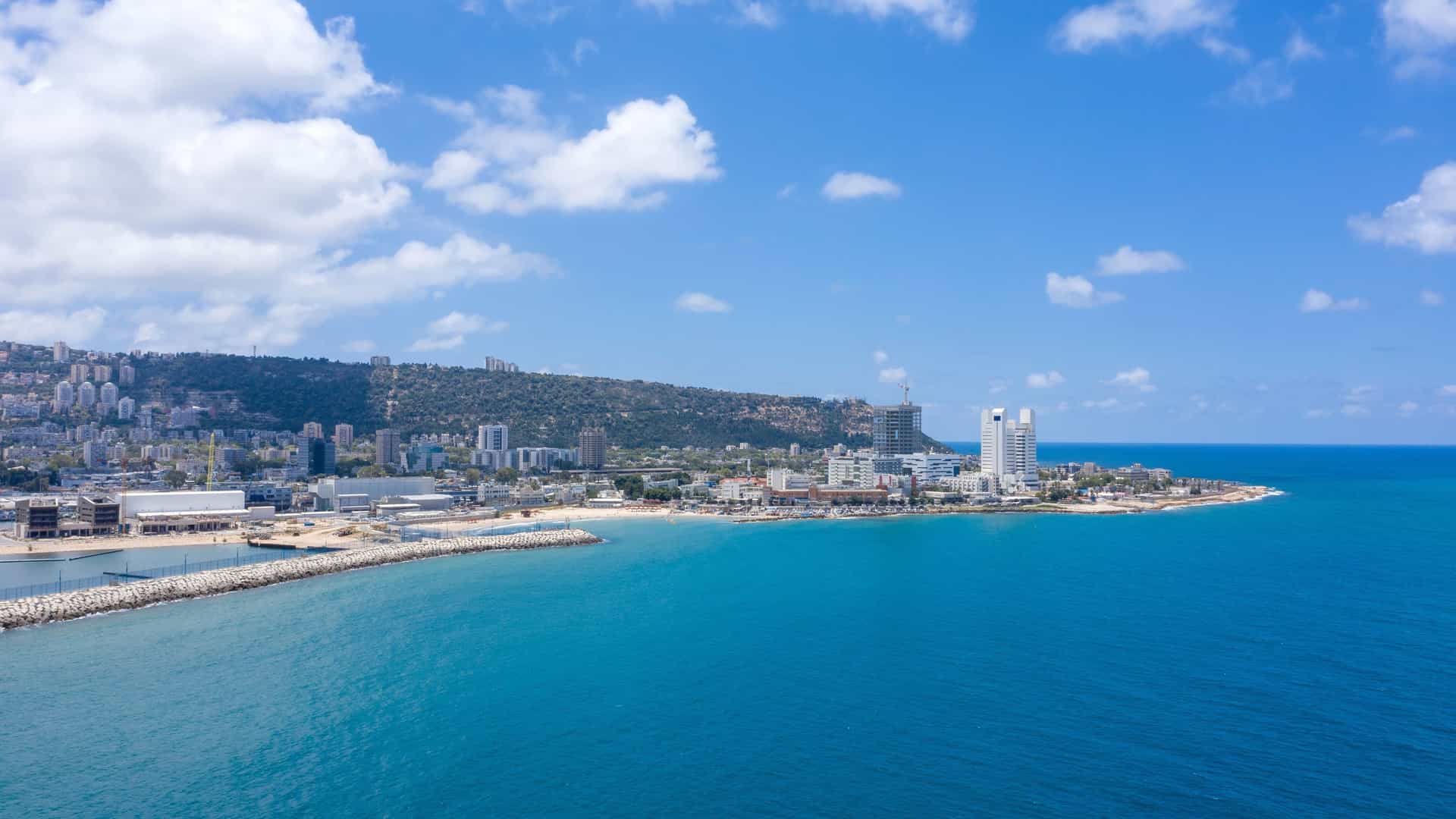  An aerial view of the Haifa Bay skyline, showcasing the bustling port with its cargo ships and cranes, and the city's downtown area. The Mediterranean Sea stretches across the foreground and a clear sky with scattered clouds is in the background.