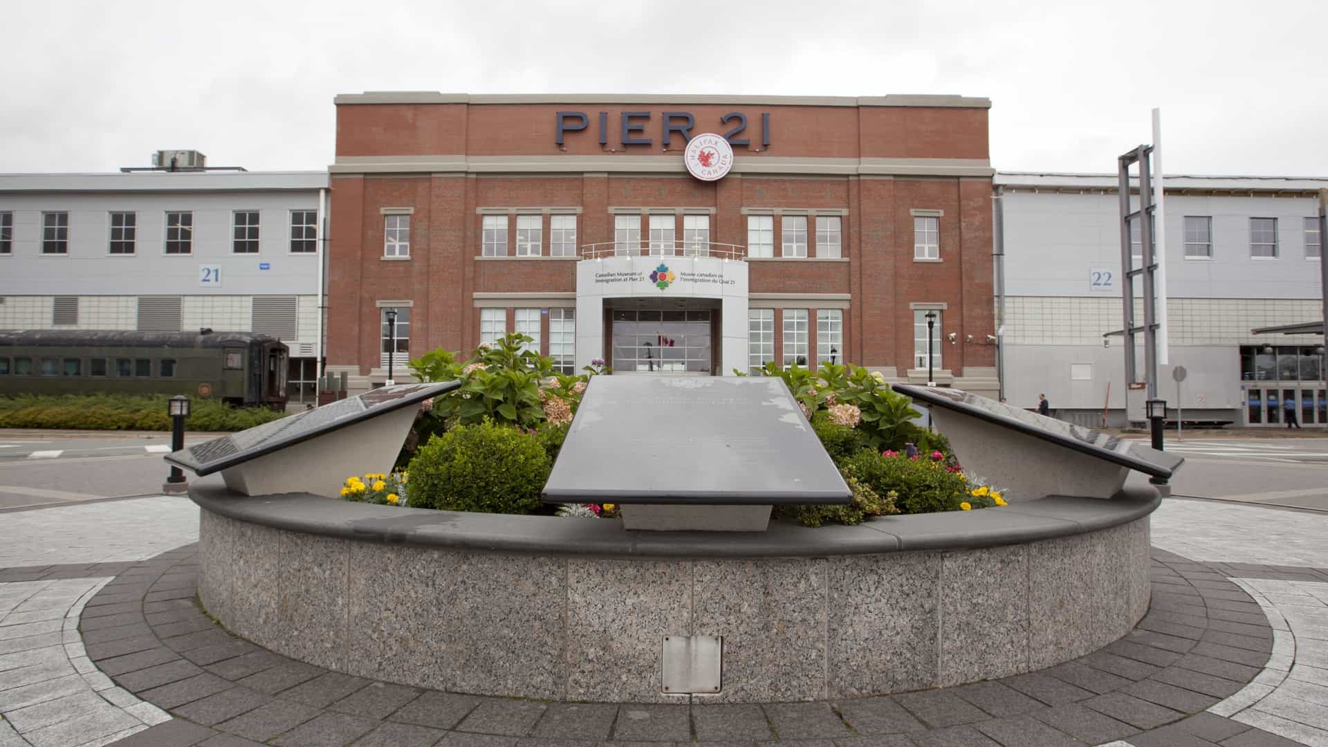 A daylight view of the exterior of the Canadian Museum of Immigration at Pier 21 in Halifax, Nova Scotia. The red brick, industrial-style building stands prominently on the waterfront cruise ship terminal, with large windows and clear signage identifying it as Canada's national museum of immigration.