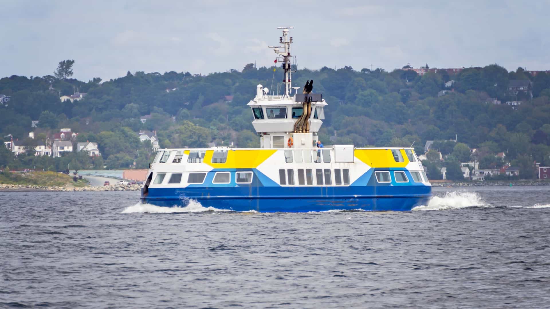 A classic orange and white Halifax Transit ferry glides across the water of Halifax Harbour in Nova Scotia, leaving a wake behind it. The image captures the "seaside commute," with the modern buildings of the Halifax waterfront and the city skyline visible in the distance under a blue sky.