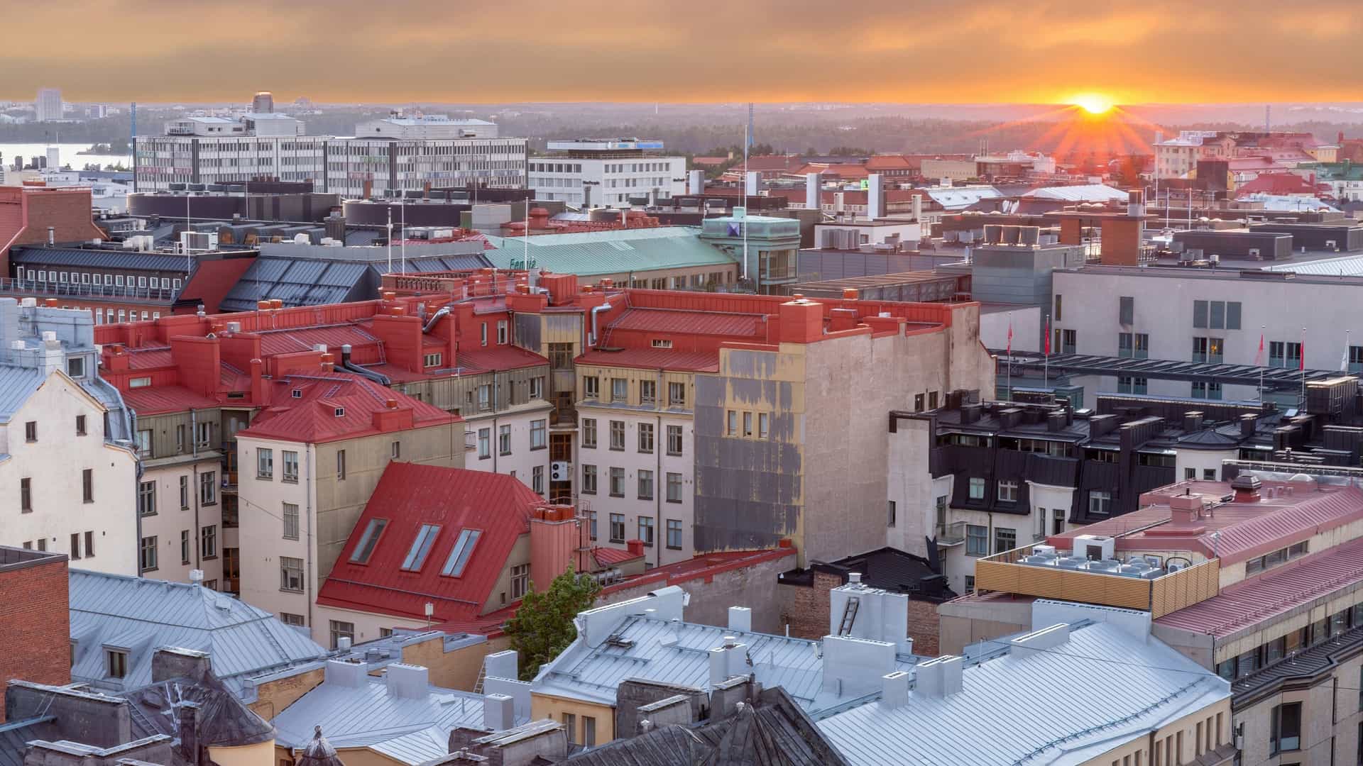 A row of distinctive, brightly colored Art Nouveau (Jugendstil) residential buildings in the Design District of Helsinki, Finland. The buildings are painted in alternating shades of striking yellow, soft green, and pink, and feature multiple bay windows, small balconies, and tall, dark, pointed turret roofs against a blue sky with white clouds. Several cars are parked along the street in front of the facades.