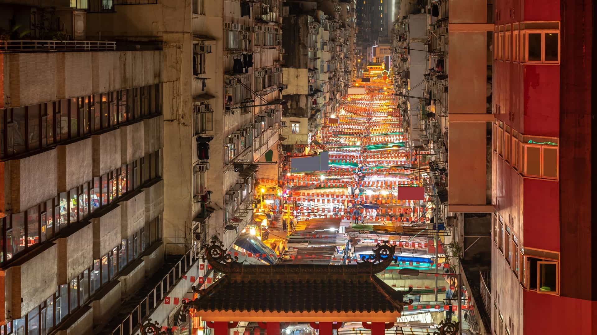 An aerial or high-angle view of the vibrant and illuminated Temple Street Night Market in Kowloon, Hong Kong, captured at night. The image shows the market stalls packed onto the street, bright lighting, and crowds of people walking and shopping in the famous cultural attraction.
