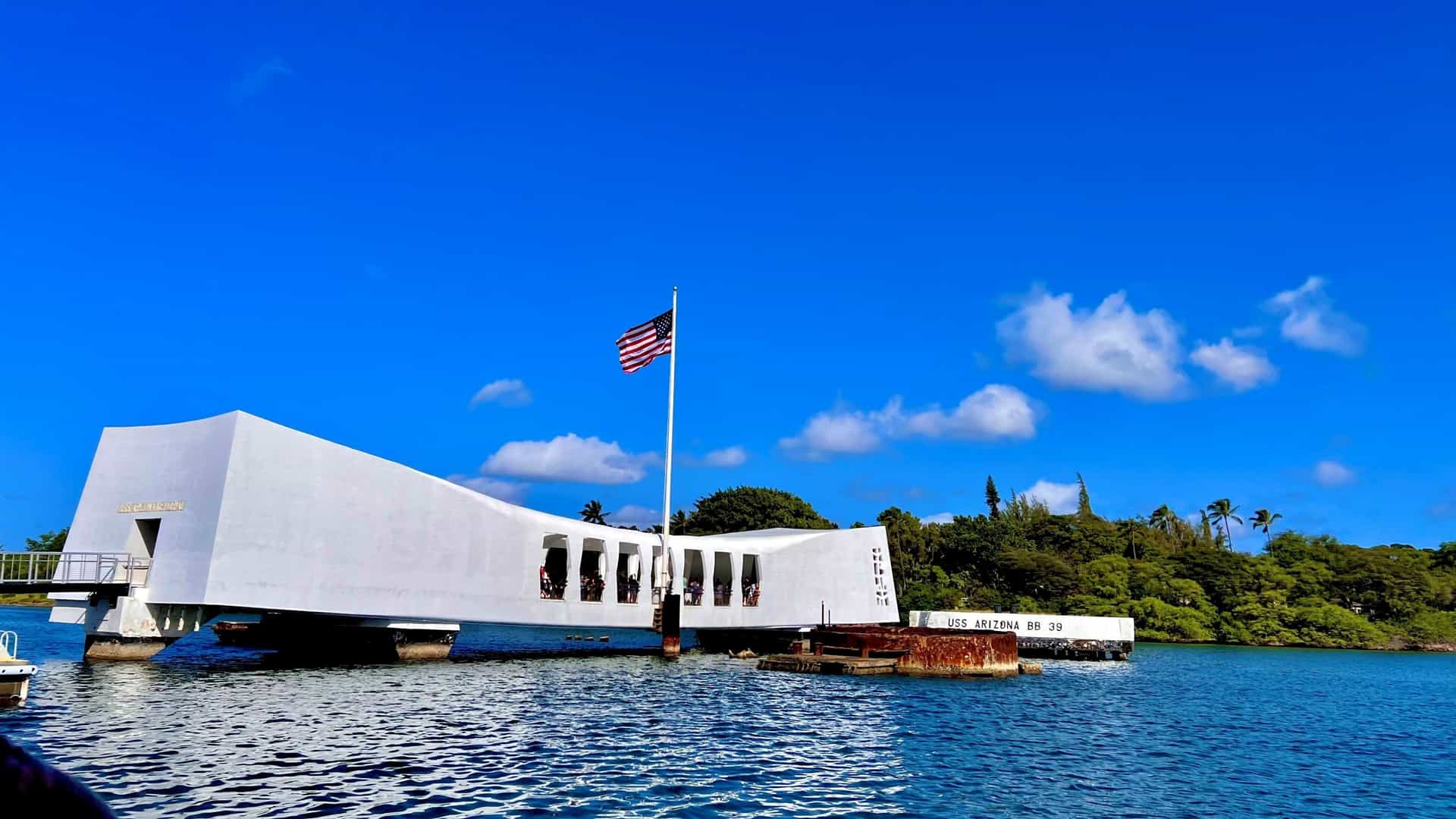A view of the USS Arizona Memorial at Pearl Harbor, Oahu, Hawaii. The distinct, white, sloping structure is visible, spanning the sunken hull of the USS Arizona battleship. The image is taken over the water of the harbor, commemorating the servicemen killed during the attack on Pearl Harbor on December 7, 1941.