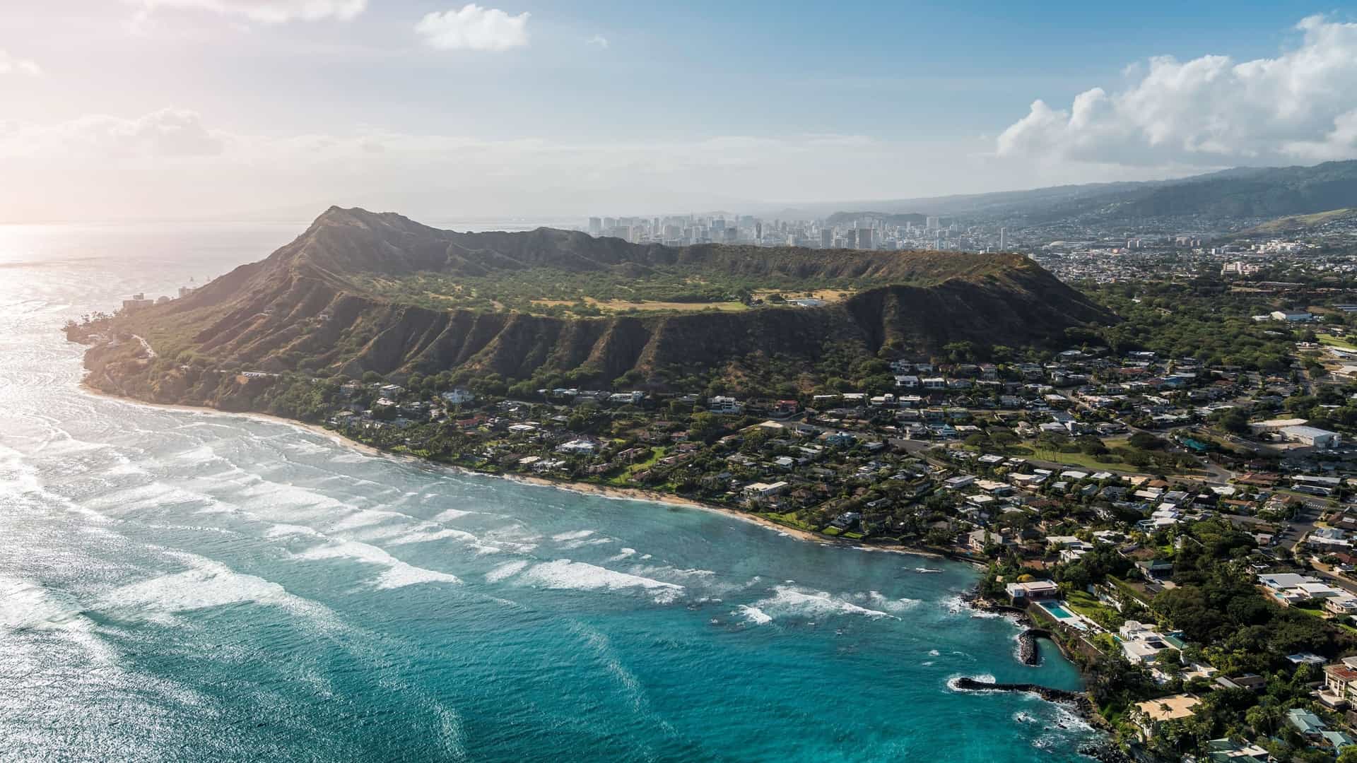  An aerial view of Diamond Head, a volcanic tuff cone, on the island of O'ahu, Hawaii. The image shows the distinct crater shape and its steep, green slopes. The photo captures the contrast between the natural landmark and the surrounding urban landscape of Honolulu, with city buildings and Waikiki beach visible in the background.
