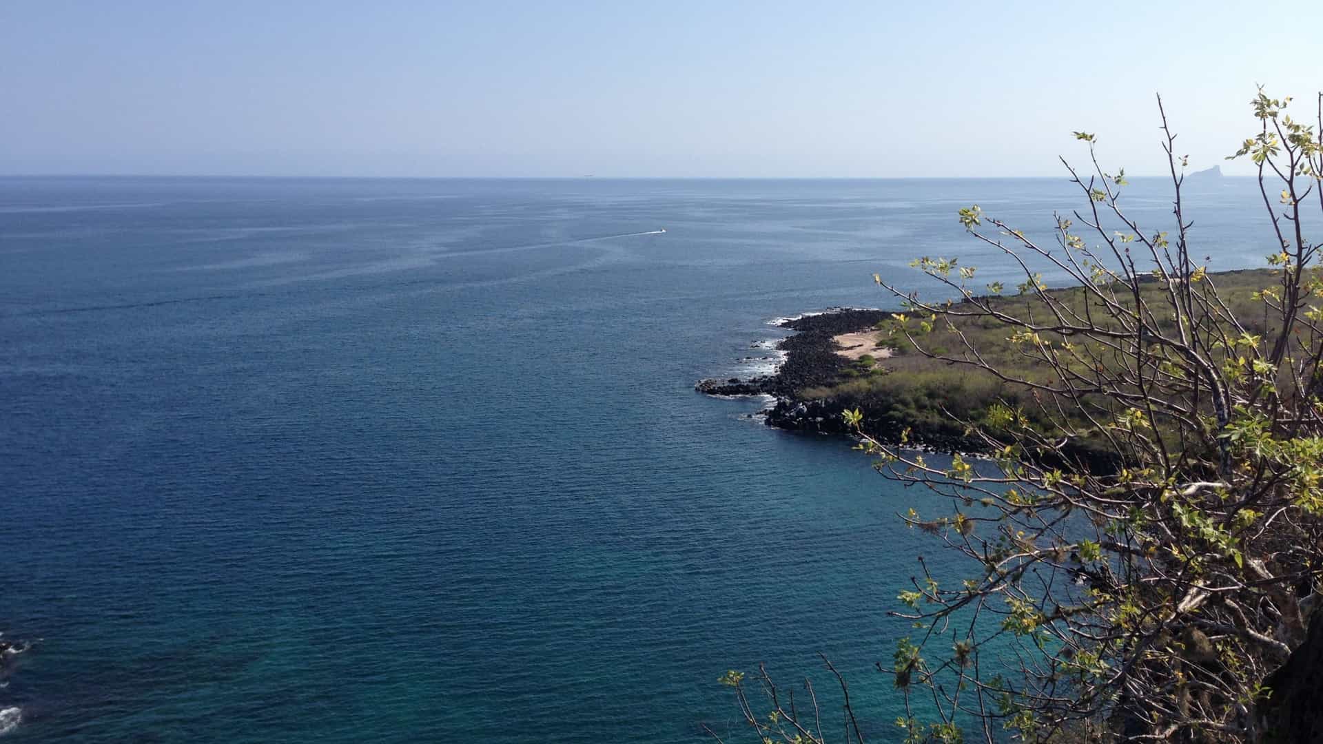 A wide, panoramic view from the Mirador Cerro Tijeretas on San Cristóbal Island in the Galápagos, looking out over the coastline and ocean. The elevated view shows a rugged, rocky shoreline and a calm bay of dark blue water, with the famous Kicker Rock (León Dormido) visible as a distinct, dramatic rock formation rising from the sea in the distant background.