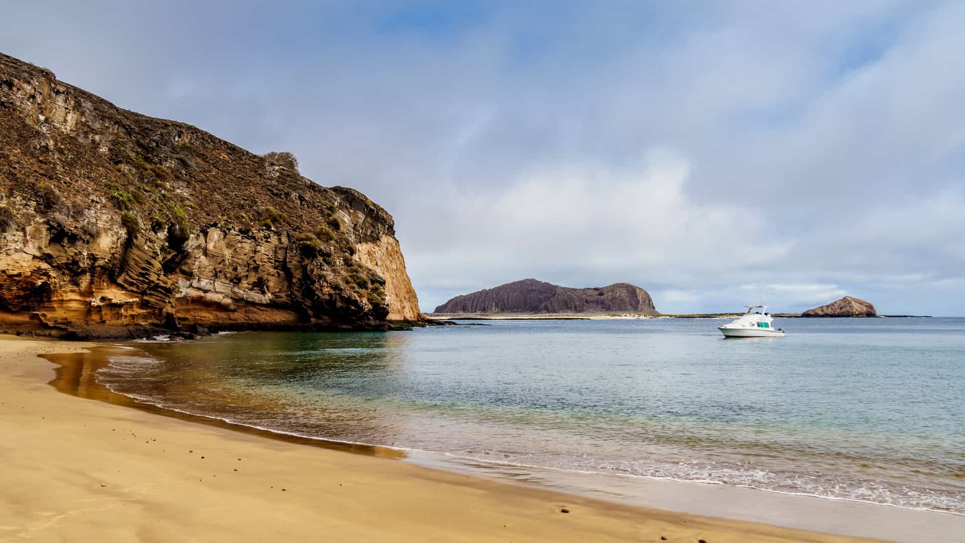 A wide, sunny view of a small, rocky beach at Punta Pitt, San Cristóbal Island, in the Galápagos. The foreground shows dark sand and volcanic rock with a gentle turquoise ocean lapping at the shore, backed by the distinctive reddish-brown cliffs and sparse, dry coastal vegetation of the volcanic landscape.