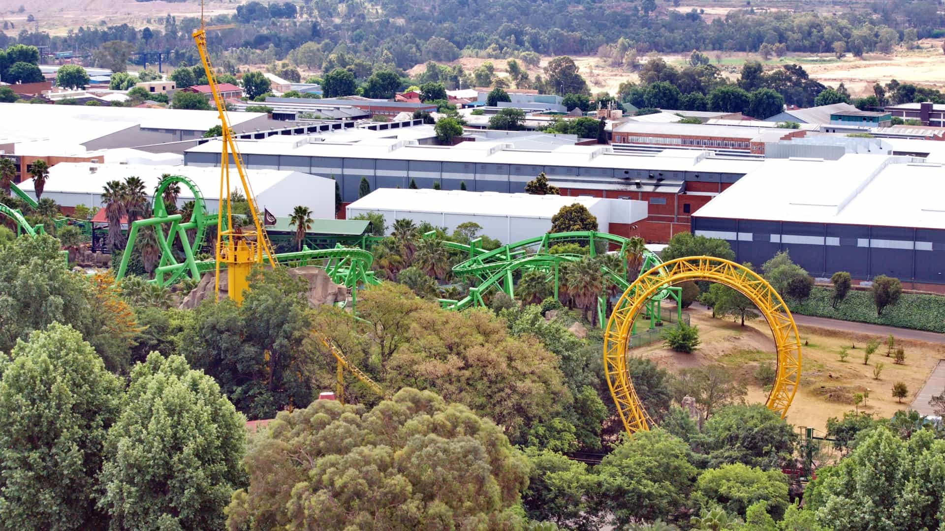 An elevated, wide-angle view of the Gold Reef City Theme Park in Johannesburg, South Africa. The foreground is dominated by the bright yellow, vertical loop of the Golden Loop roller coaster and the twisting green track of the Anaconda inverted coaster, nestled among dense green trees. White industrial and commercial buildings, along with a dry, dusty landscape, stretch into the background.