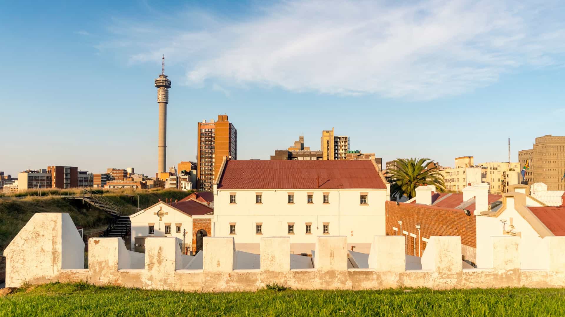 A view within the Constitution Hill Human Rights Precinct in Johannesburg, capturing the historic architecture of the Old Fort Prison. The image features white-walled, low-slung buildings from the former prison complex with exposed brick details, set against a paved courtyard and a clear blue sky. In the background, the prominent, cylindrical Telkom Tower of the Johannesburg skyline is visible, emphasizing the site's central location in the city.