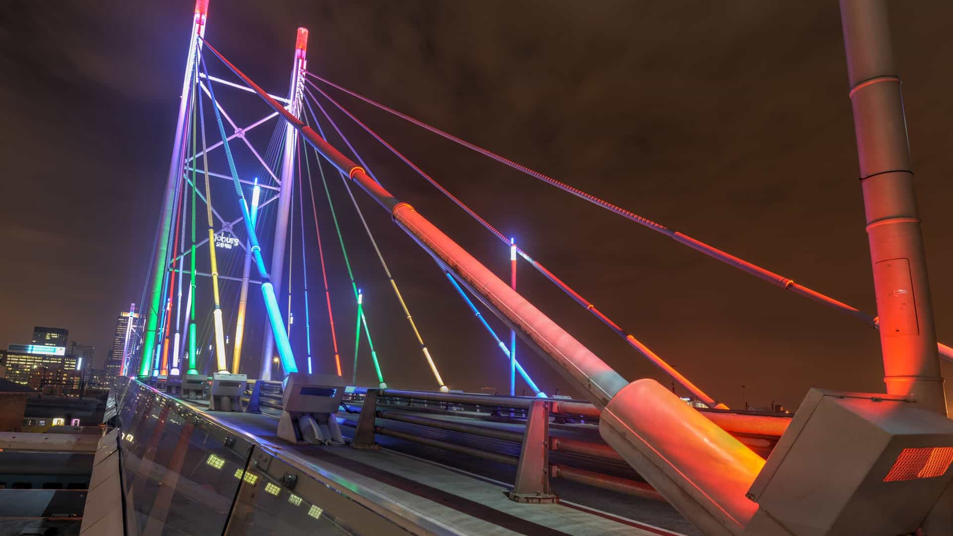 A dramatic, low-angle night photograph of the Nelson Mandela Bridge in Johannesburg. The white, cable-stayed bridge is brightly illuminated by warm yellow lights, contrasting with the dark night sky. The bridge's main deck extends across the frame, suspended by cables that connect to the tall central pylon. Streetlights glow along the road, and the background shows the soft, blurred lights of the surrounding cityscape.