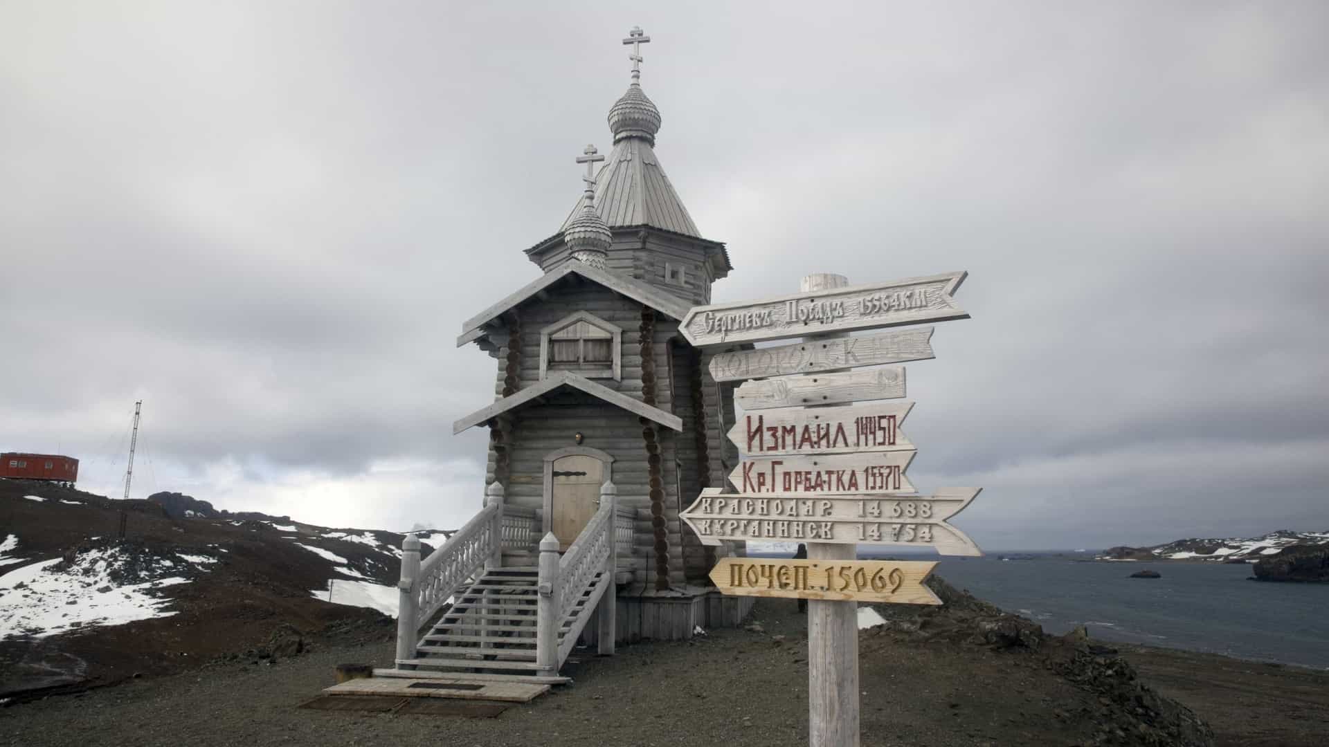A tall, wooden Russian Orthodox church, known as Holy Trinity Church, perched on a rocky, snow-dusted hill overlooking the coastline of King George Island, Antarctica. The structure is built in a traditional Russian style, standing 15 meters high, and is constructed from Siberian pine logs, having been disassembled, shipped, and reassembled at the site near the Bellingshausen research station. The image often highlights the contrast between the traditional, elaborate wooden architecture and the remote, icy Antarctic landscape.
