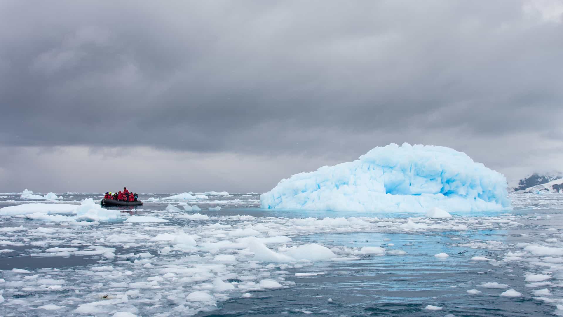 A low-angle view of a Zodiac inflatable expedition boat (or similar Rigid Inflatable Boat) moving slowly through a dense field of brash ice and small icebergs (or bergy bits) on the open sea. The water is a dark, cold gray or blue, partially obscured by countless small, white and blue chunks of ice. The Zodiac is typically carrying passengers wearing brightly colored, insulated, waterproof gear (such as red or yellow parkas), who are seated along the perimeter of the boat, gazing at the surrounding ice formations and vast, rugged polar landscape.