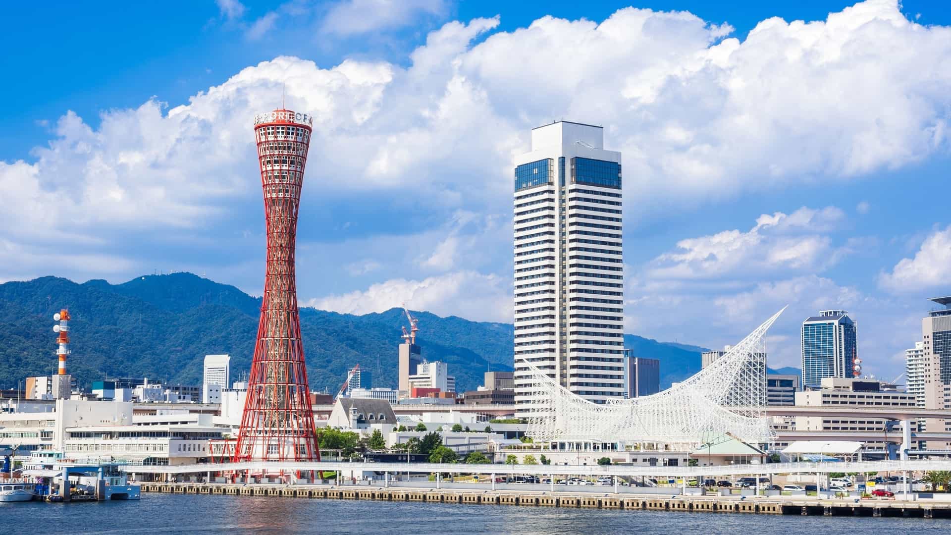 A daytime wide shot of the Kobe waterfront in Japan, featuring the iconic red lattice Kobe Port Tower and the large white, wave-like roof of the Kobe Maritime Museum in Meriken Park. The landmarks stand prominently over the harbor, with modern buildings and ships visible in the background under a blue sky.