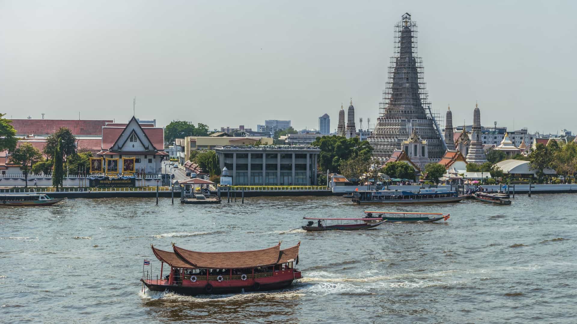  A long tail boat navigates the busy Chao Phraya River in Bangkok, Thailand. The river is a mix of brown and green water, with buildings and modern high rises visible on the banks.
