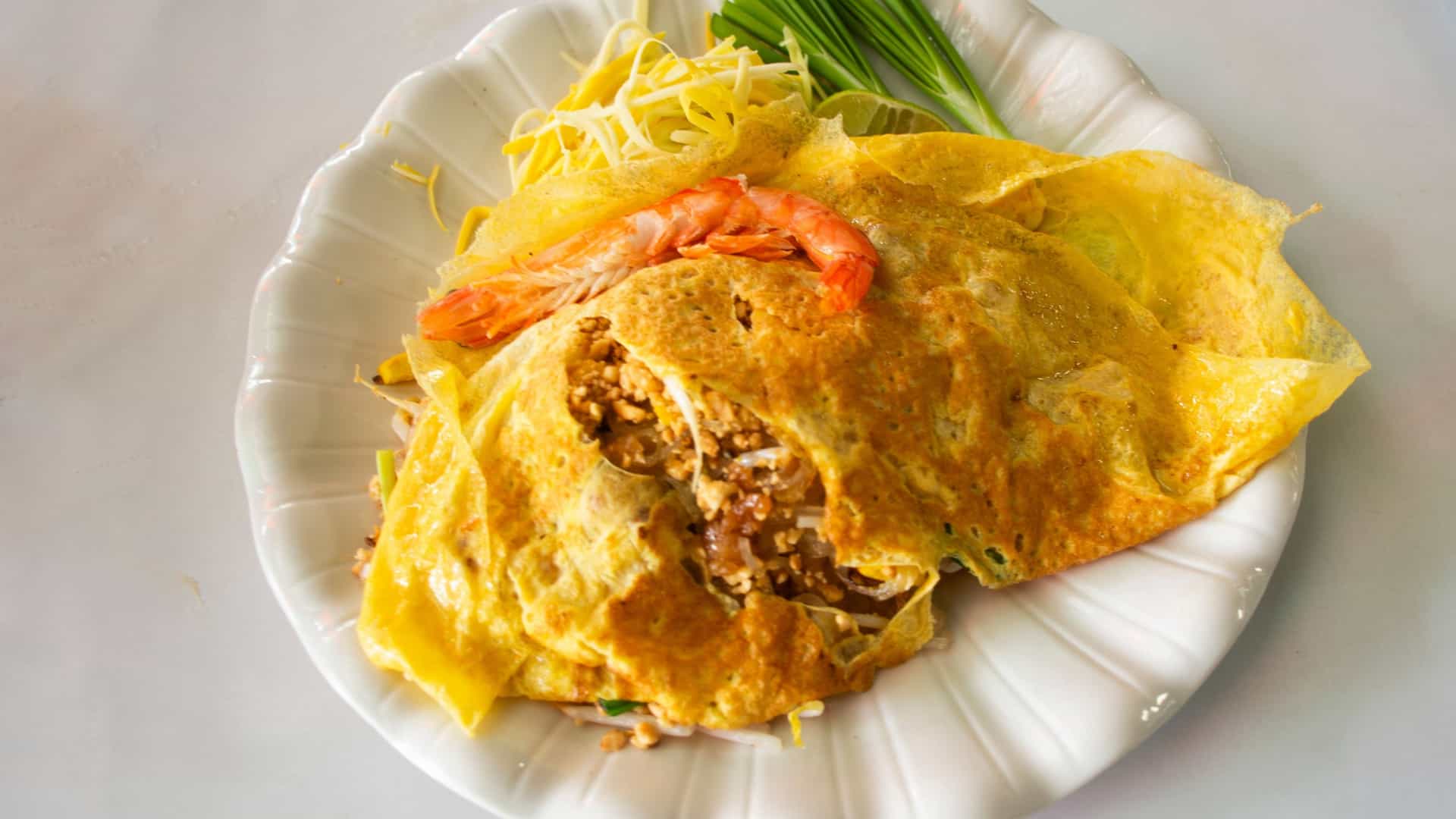  A dish of traditional Thai Pad Thai street food is served in a bowl, featuring stir fried rice noodles with a variety of ingredients. The dish is topped with crushed peanuts and fresh lime wedges on the side.