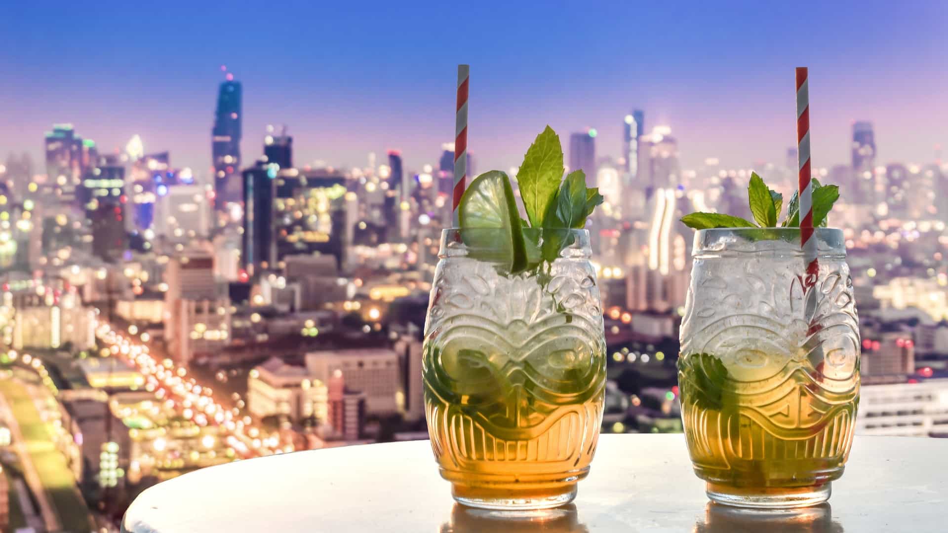A close-up view of two Mojito cocktails in tall glasses placed on a bar or table at a rooftop bar. The glasses are garnished with fresh mint and lime wedges. The background is a stunning, blurred cityscape view, likely during sunset or evening, with the lights of buildings and the urban skyline visible below.