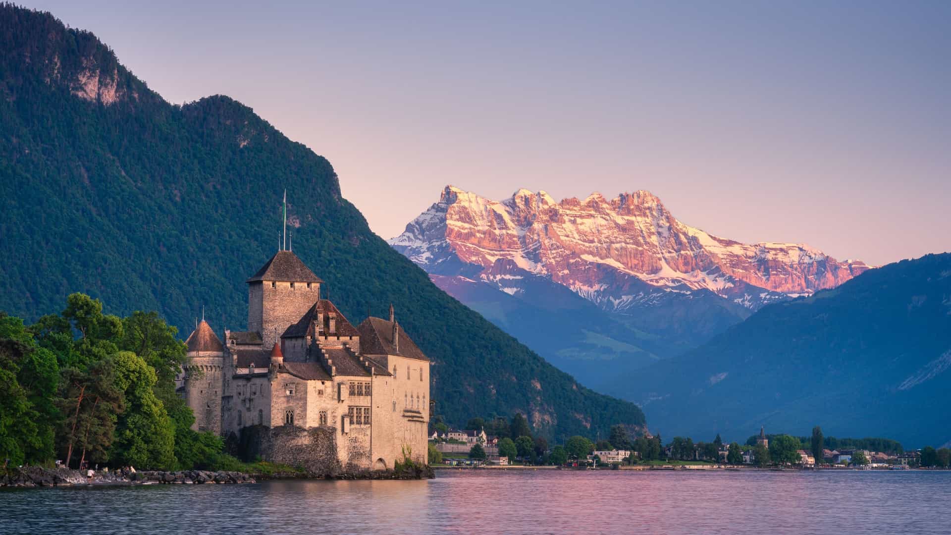 A beautiful, wide-angle shot of the Château de Chillon in Montreux, Switzerland, at sunset or blue hour. The medieval castle, which sits on a rock on the edge of Lake Geneva (Lac Léman), is illuminated against the dramatic sky. The calm water of the alpine lake reflects the castle and the warm colors of the fading light, with the dark outlines of the surrounding mountains visible in the background.
