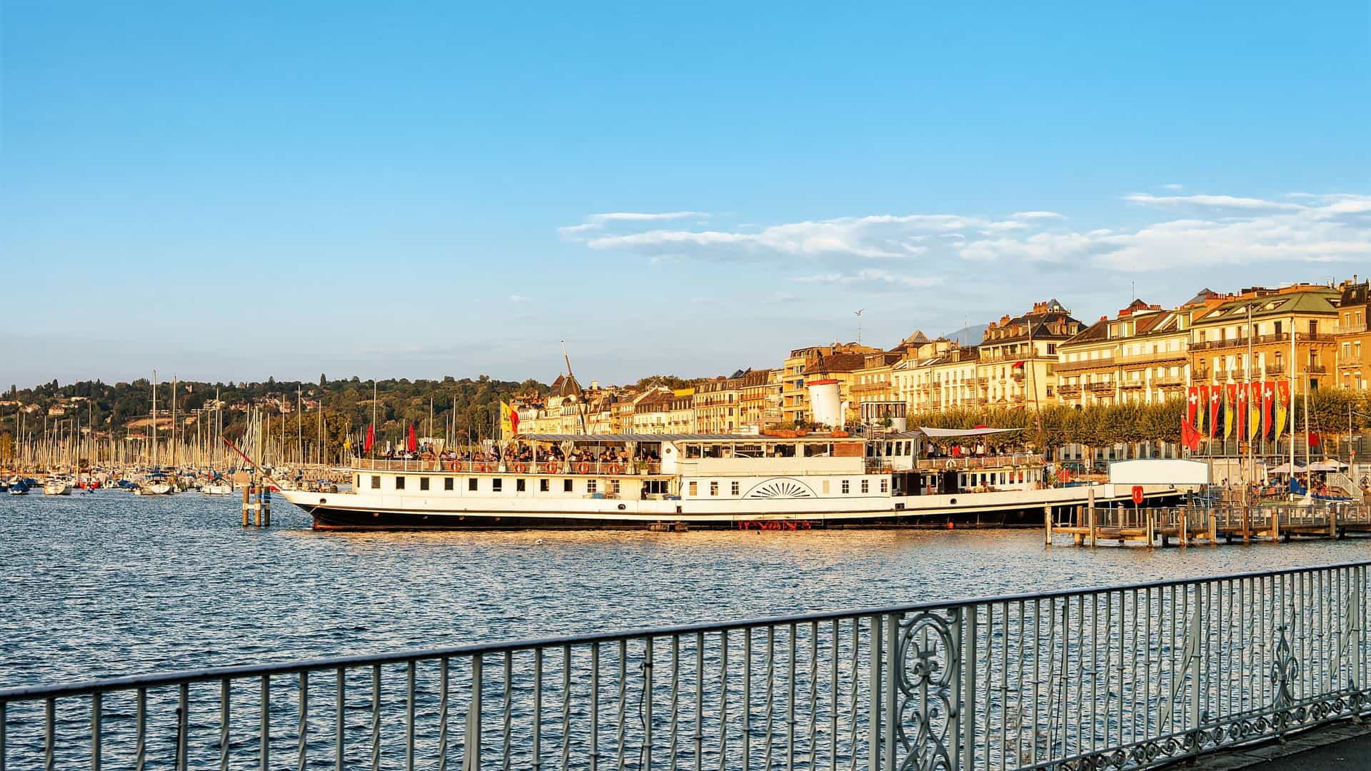  A traditional steamboat, or paddle steamer, is docked at a pier on Lake Geneva in Switzerland. The image is taken from a distance, showing the boat and pier in the foreground with the calm lake and mountains in the background under a blue sky.