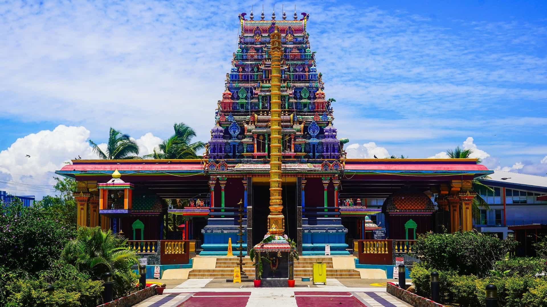 A colorful, wide-angle shot of the Sri Krishna Kaliya Temple in Lautoka, Fiji. The main structure features a towering, multi-tiered pyramidal temple tower (gopuram or vimana) covered in intricate, polychromatic carvings of Hindu deities. A prominent golden pillar (dhvajastambha) stands on a raised platform in front of the entrance, all set beneath a bright blue sky with white clouds.