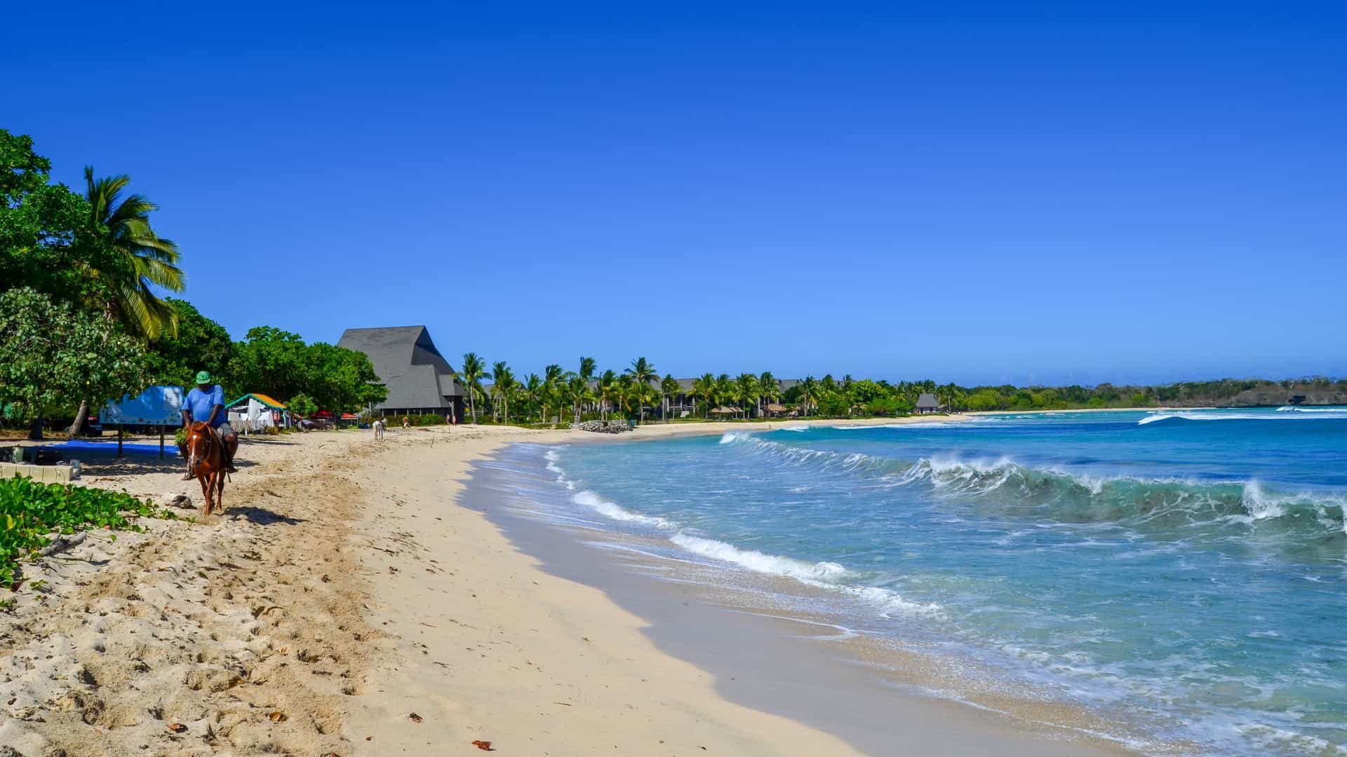 A panoramic view of the popular Natadola Beach on Viti Levu Island, Fiji. The image captures a wide, crescent-shaped sandy beach fringed by lush green vegetation and palm trees. The turquoise ocean water is clear and calm, with gentle waves breaking near the shore under a bright blue sky dotted with white clouds.