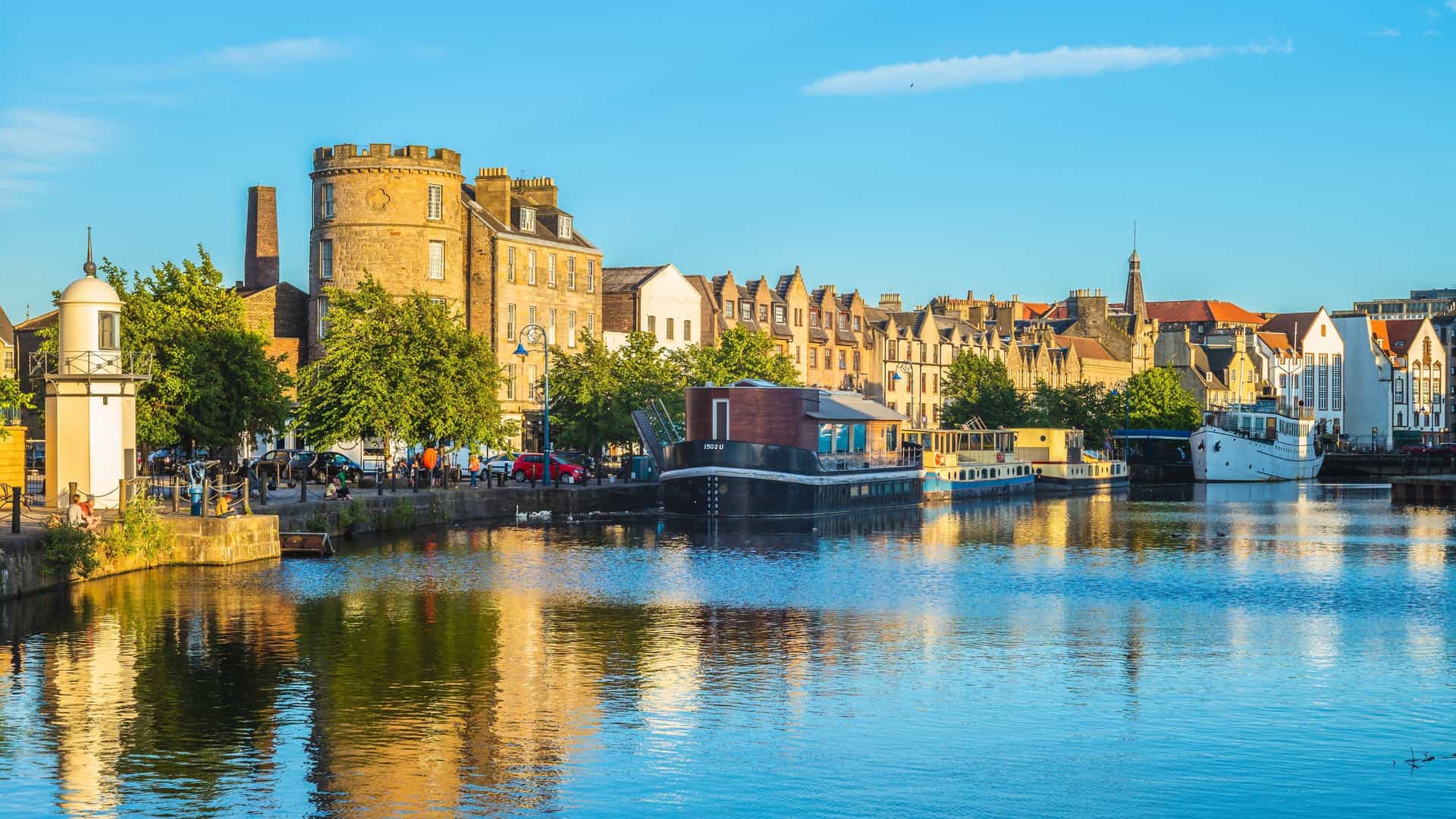 A scenic view of The Shore area in Leith, Edinburgh, UK, where the Water of Leith meets the docks. The image features historic and modern buildings lining the cobbled quayside, with various small boats and yachts moored in the water. This picturesque area is known for its waterfront restaurants and proximity to major Leith attractions.