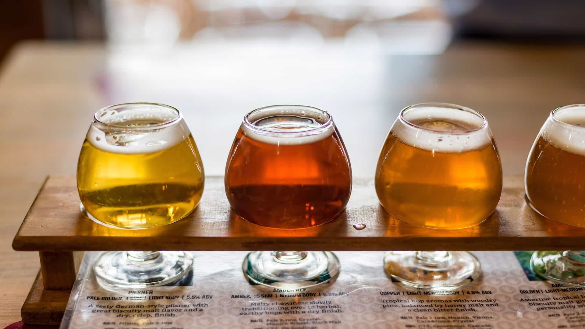 A still-life photograph featuring a wooden flight paddle holding four small, footed glasses of liquids, likely a tasting selection from a local microbrewery. The contents vary in color from pale yellow to rich amber. Beneath the paddle, a printed menu describes the characteristics of the different beverages.