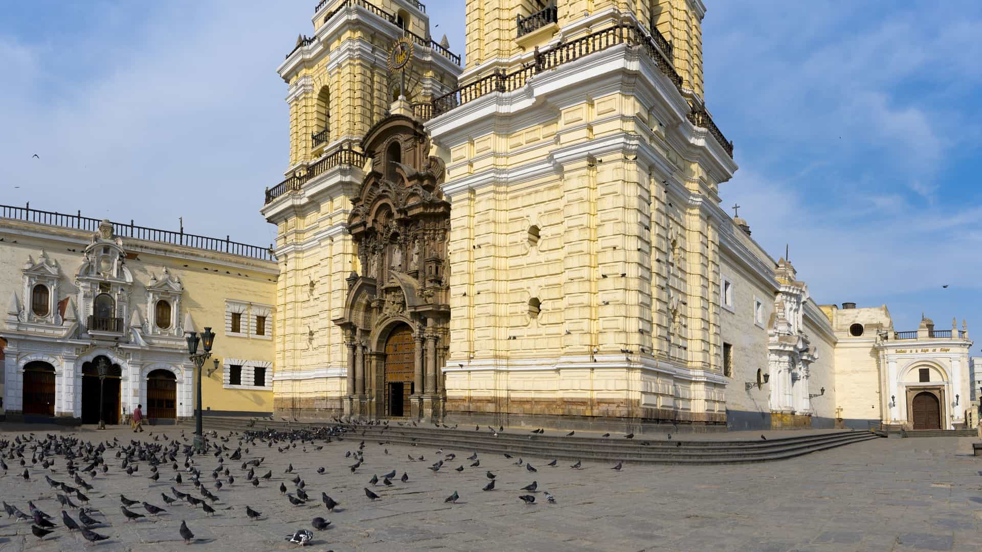A low-angle, wide view of the Basilica and Convent of San Francisco in Lima, Peru. The large, yellow-colored Baroque church features two prominent bell towers and an intricately carved dark stone portal. A large flock of pigeons is gathered on the stone plaza in the foreground, with the adjacent convent buildings visible to the left and right under a bright blue sky.