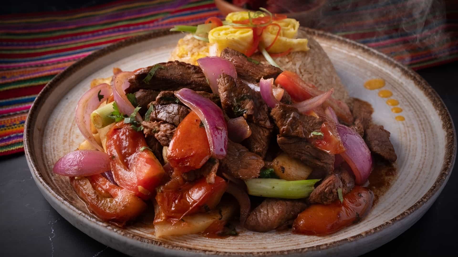 A close-up, top-down view of a classic Peruvian dish called Lomo Saltado. The stir-fry consists of thick-cut strips of seasoned beef tenderloin, slices of red onion, and wedges of tomato, all tossed in a dark, rich sauce and garnished with fresh chopped cilantro or parsley. It is accompanied by a generous portion of golden-brown French fries and a side of fluffy white rice, characteristic of how the dish is traditionally served.