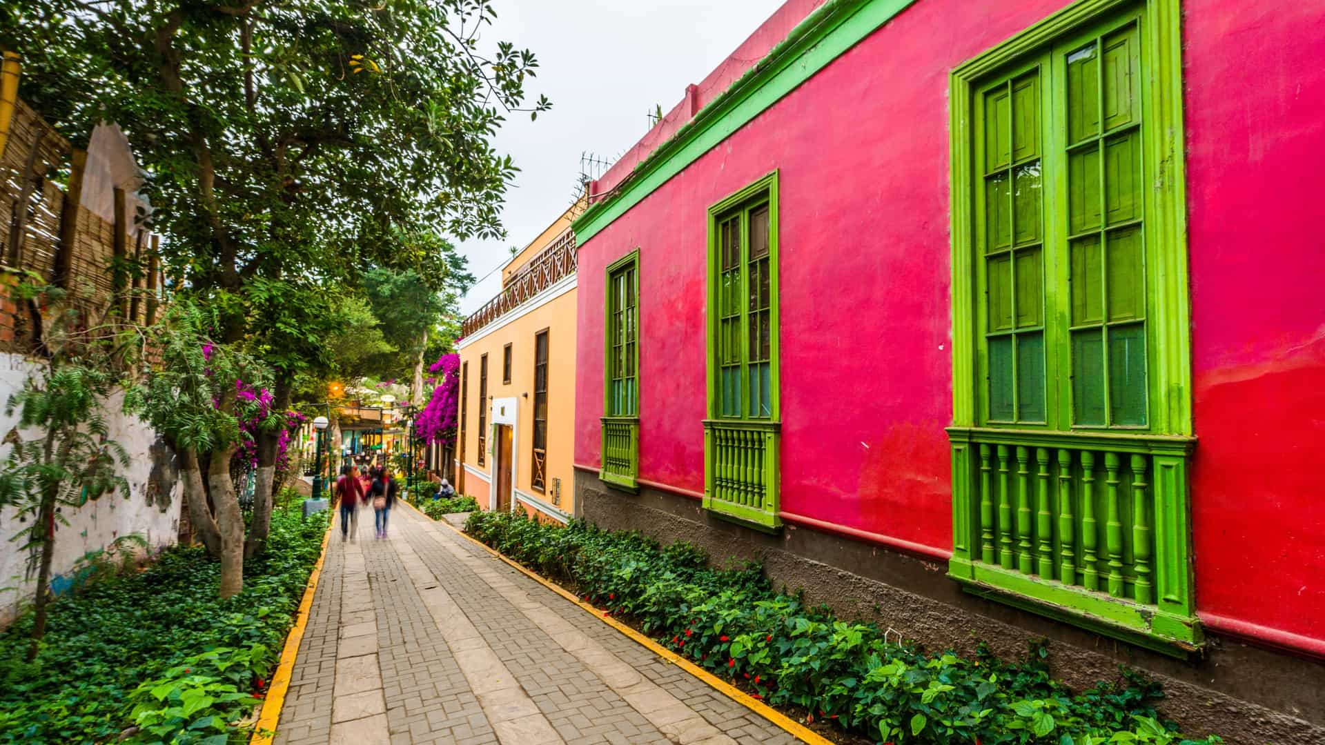 A view of the historic Barranco district in Lima, Peru, featuring a colorful street with colonial-style buildings painted in vibrant shades of red and blue, leading toward the ocean under a bright sky.