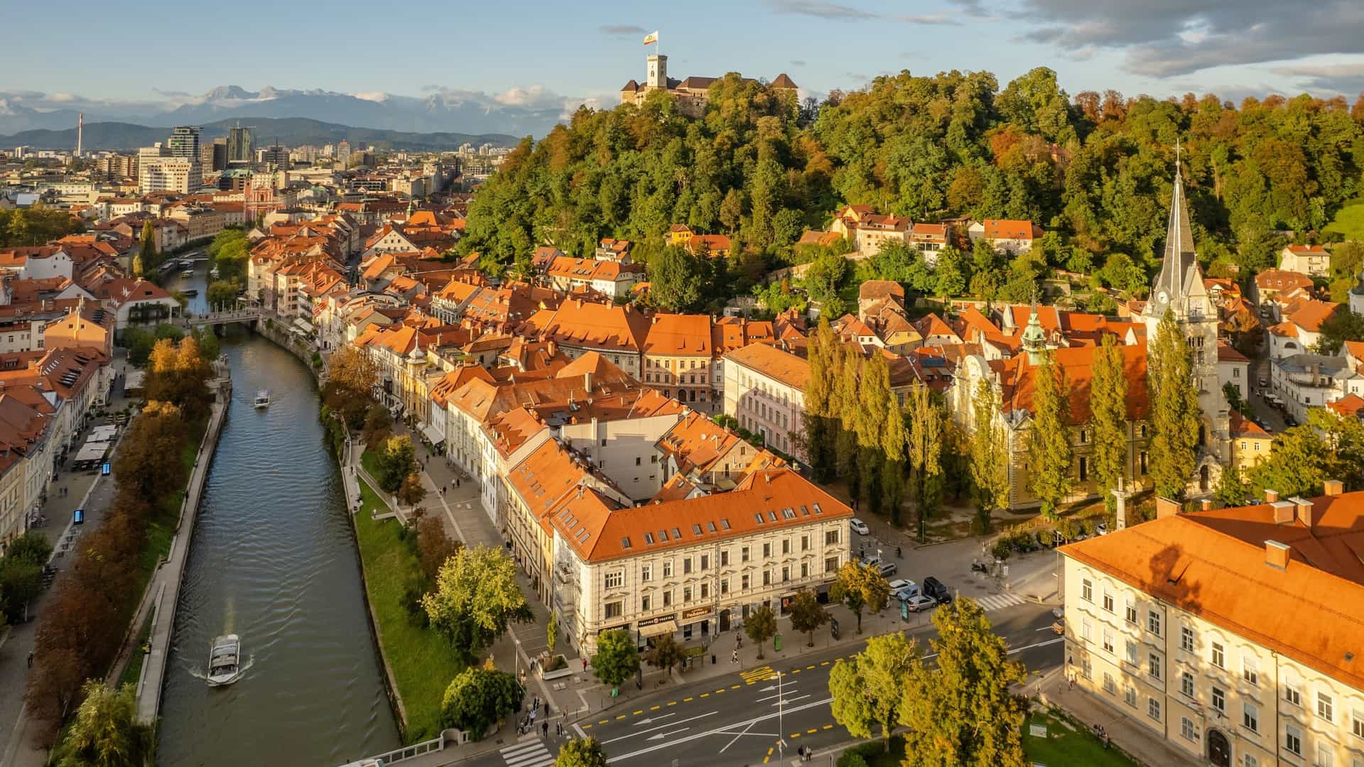  An aerial view of Ljubljana Old Town in Slovenia on a sunny day. The image shows the red rooftops of the historic buildings, the curving Ljubljanica River, and the Ljubljana Castle perched on a hill. A mountain range is visible in the background.