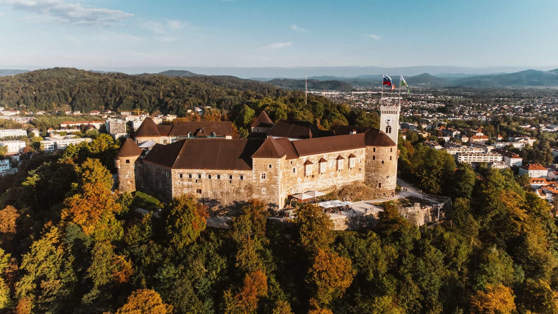  An aerial view of Ljubljana Castle, a medieval fortress perched on a hill above the city of Ljubljana, Slovenia. The image showcases the castle's fortified walls, towers, and courtyards, with the city's red rooftops and surrounding greenery visible in the background.
