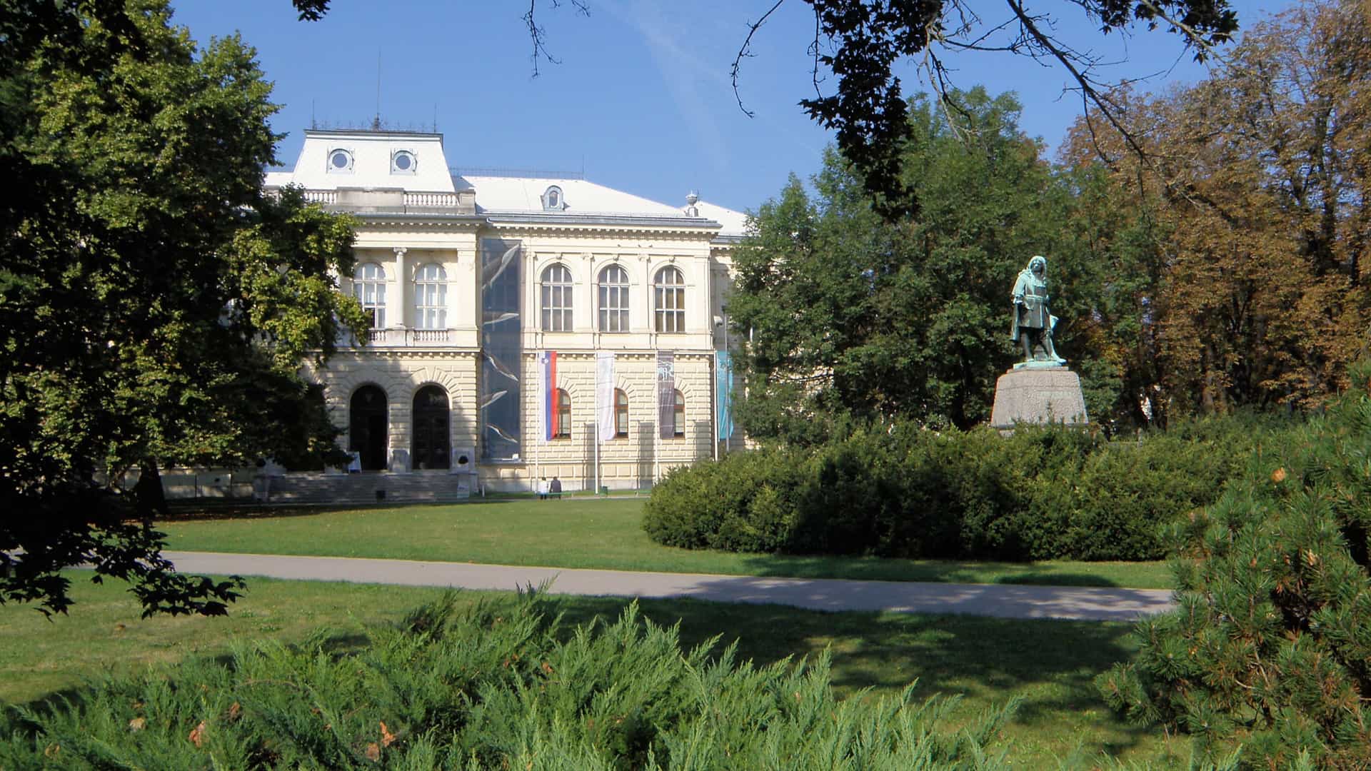 A bright, daytime photograph of a neoclassical or Neo-Renaissance museum building, likely the National Museum of Slovenia in Ljubljana, seen from across a grassy park. The light-colored, multi-story building features arched windows and a prominent entrance. In the foreground, a large bronze statue on a pedestal stands amidst green bushes and trees, with the rest of the park lawn filling the lower part of the frame.