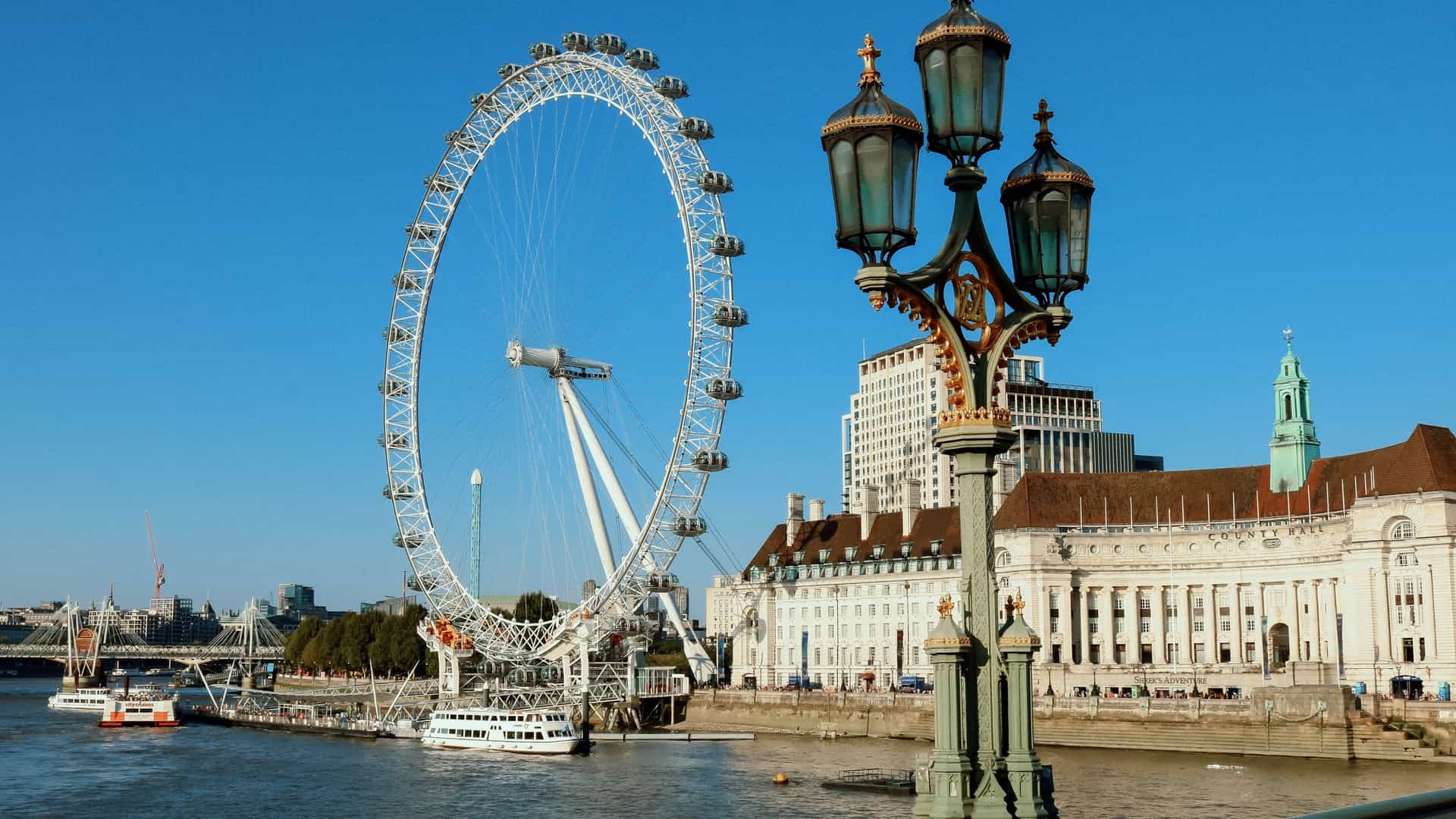 A prominent view of the London Eye, a massive observation wheel, seen from the Westminster Bridge overlooking the River Thames in London. The Ferris wheel's spoke structure and passenger capsules are clearly visible, dominating the skyline on the South Bank.