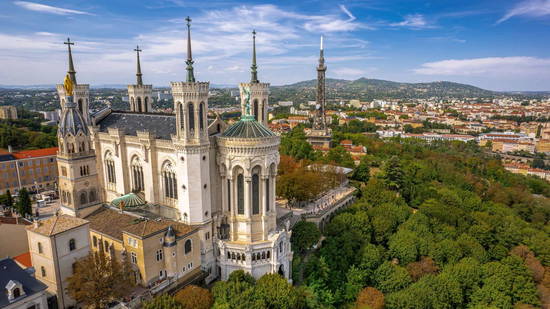 An aerial view of the Basilica of Notre Dame de Fourvière, a large, ornate Roman Catholic church with distinctive Byzantine and Romanesque architecture, located on a hilltop overlooking the city of Lyon, France. The basilica features multiple towers and a prominent dome. The dense urban landscape of Lyon stretches out in the background, with the Saône River visible winding through the city.