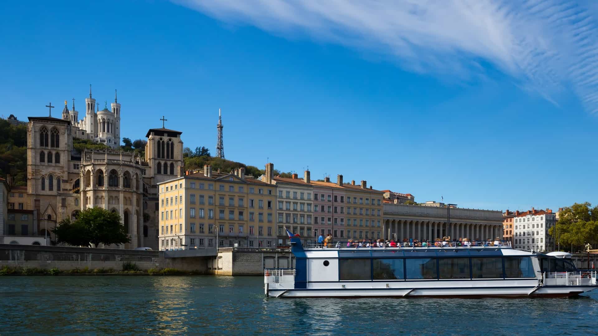 A sunny, clear-day view of the Saône River in Lyon, France, showing a large, modern tourist boat or river cruise vessel on the water in the foreground. In the background, the colorful residential buildings of the old town line the riverbank, with the historic, white spires of the Notre-Dame de Fourvière Basilica and the metallic tower standing prominently atop the hill.