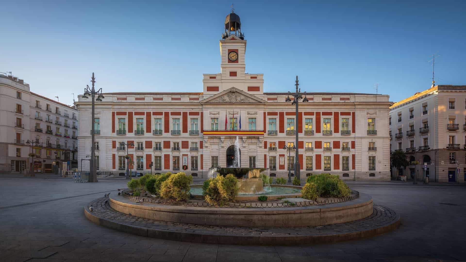 A wide-angle, daytime view of the semi-circular Puerta del Sol square in Madrid, Spain, bustling with people. The image is dominated by the historic Real Casa de Correos (House of the Post Office) building in the background, which features a prominent clock tower. In the foreground, the equestrian statue of King Charles III is visible in the center, and the square is marked by the presence of a few palm trees and the iconic, brightly-lit Tío Pepe neon sign on a nearby rooftop.