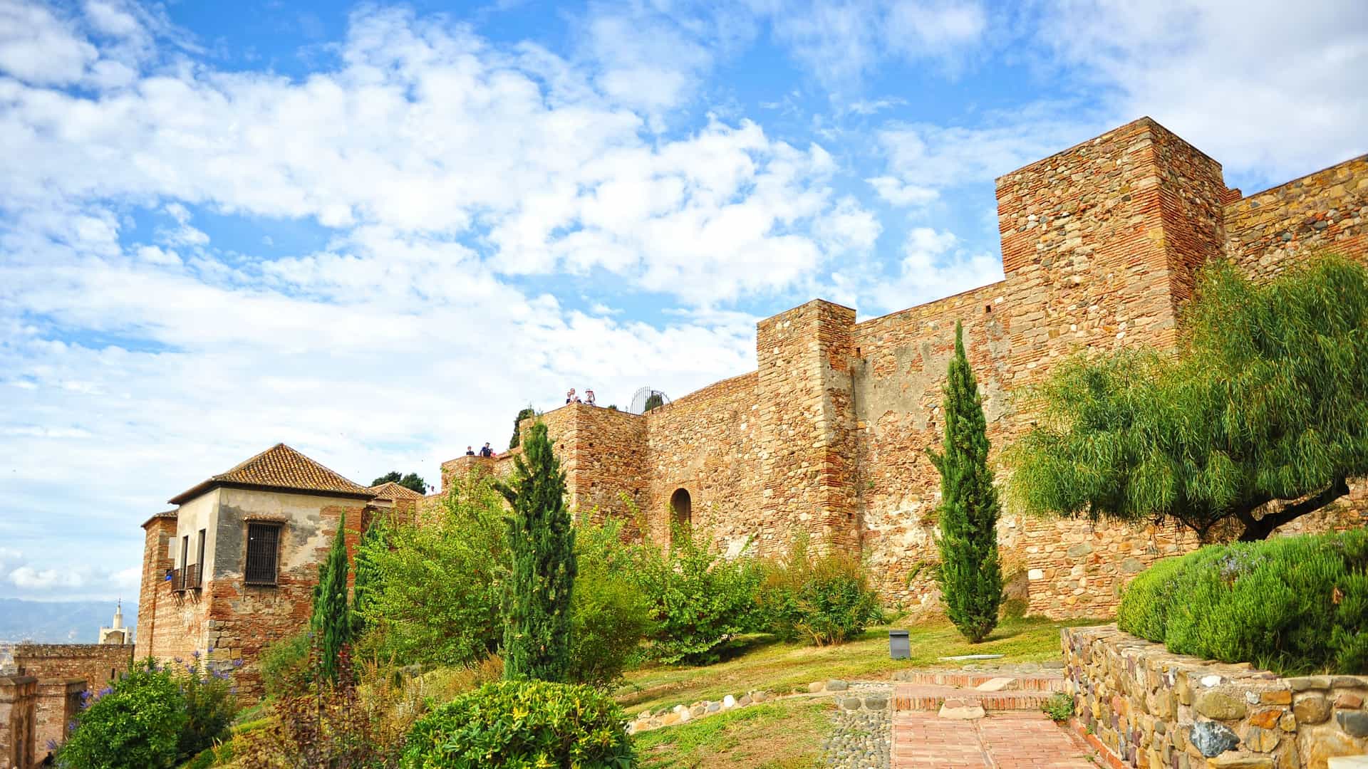 A scenic view of the Alcazaba of Málaga, a historic Moorish fortress in Málaga, Spain. The image captures the impressive architecture of the ancient fortress, with its fortified walls, towers, and lush gardens. The fortress is situated on a hill, offering a panoramic view of the surrounding city and the harbor below. The scene highlights the fortress's historical significance and its beautiful location.