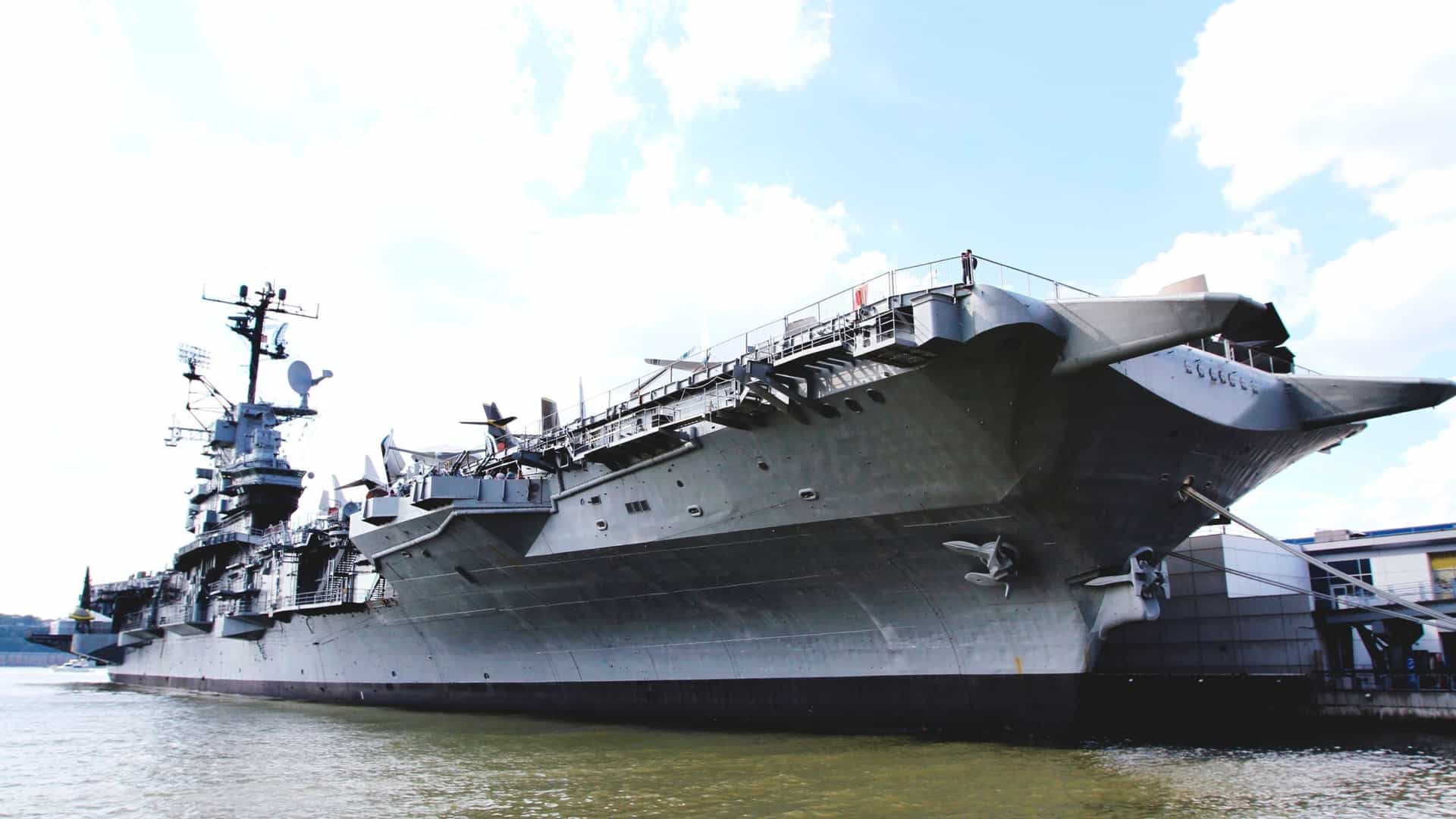 A daytime exterior view of the Intrepid Sea-Air-Space Museum in New York City. The museum is centered around the massive aircraft carrier USS Intrepid (CV-11), docked at Pier 86 on the Hudson River, with the Manhattan skyline visible in the background.