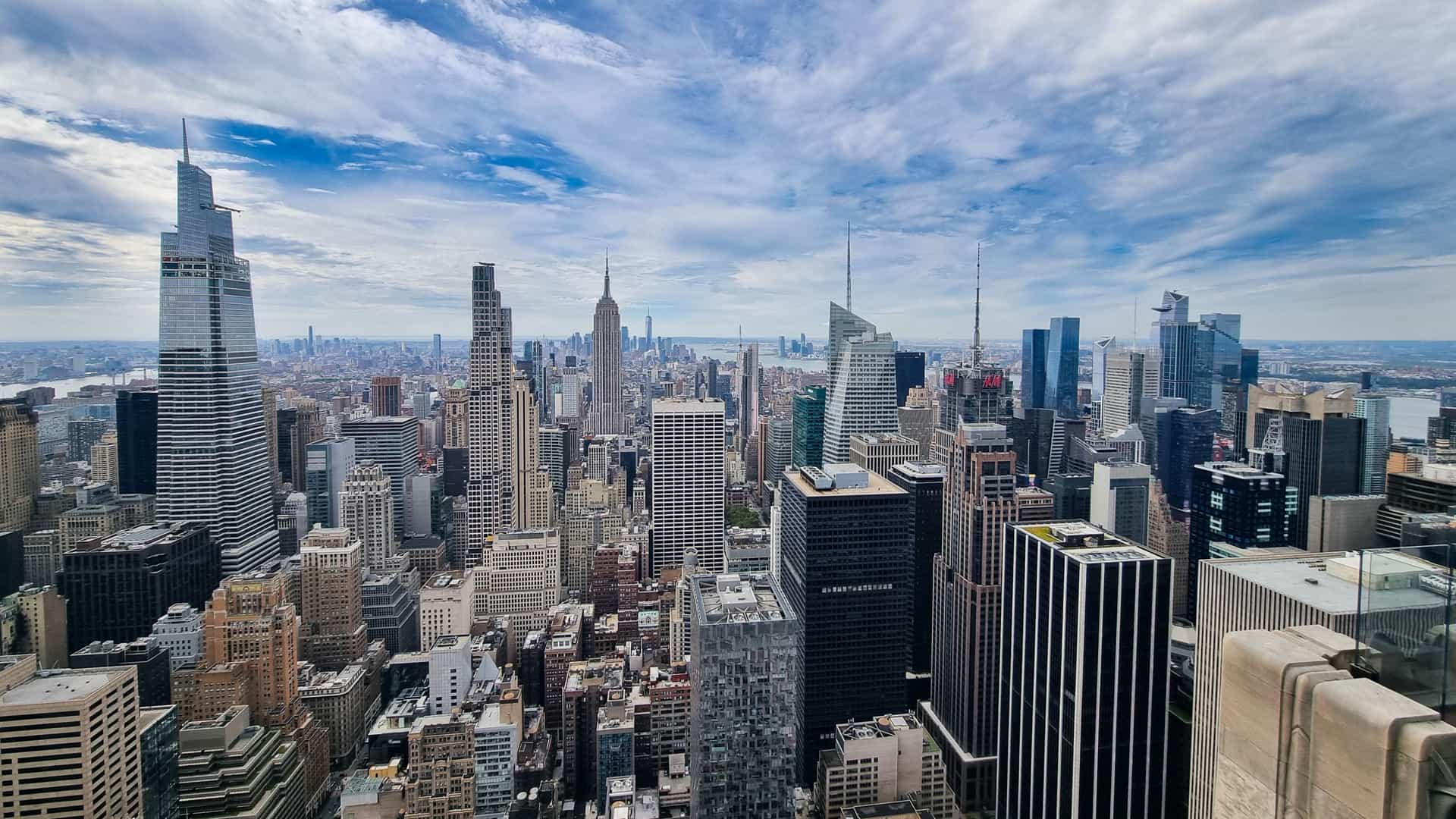 An iconic top-down view of the Midtown Manhattan skyline, seen from the Top of the Rock observation deck, showcasing the density of skyscrapers, streets, and the green expanse of Central Park in the distance.