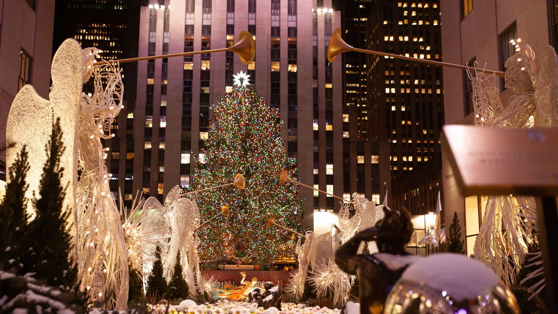 A festive night view of the Rockefeller Center Christmas Tree in New York City. The massive, brightly lit Christmas tree stands in the background, with the iconic rows of glowing golden trumpet angels (Valerie Clarebout's) lining the Channel Gardens in the foreground, creating a classic holiday scene.